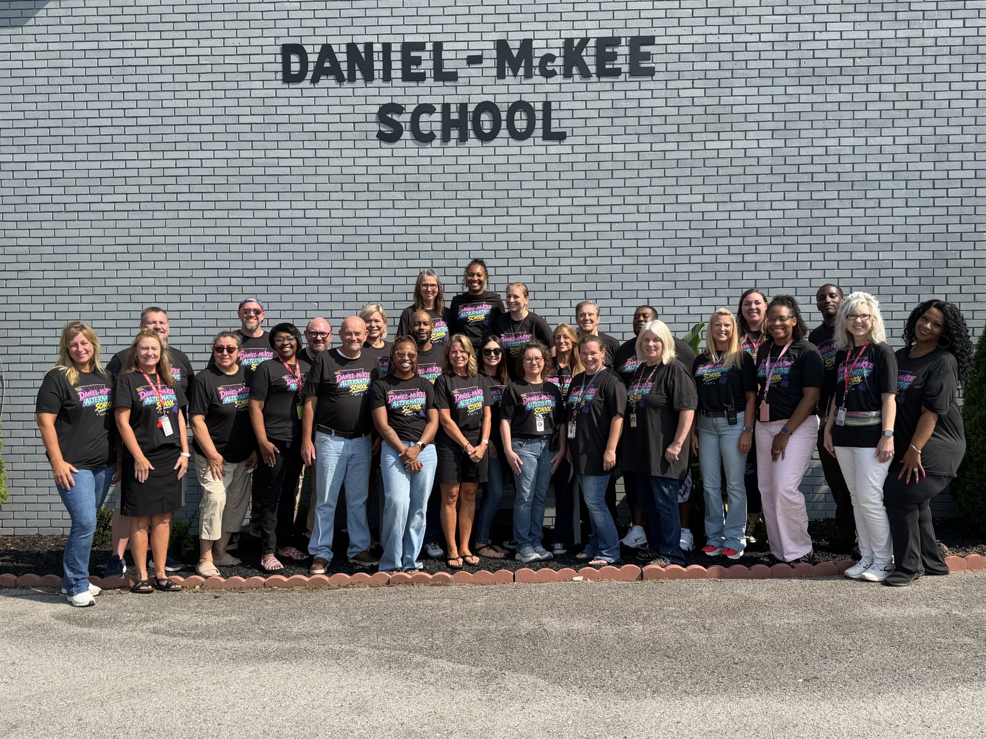 A group of adults in matching black T-shirts poses for a photo in front of Daniel-McKee School.