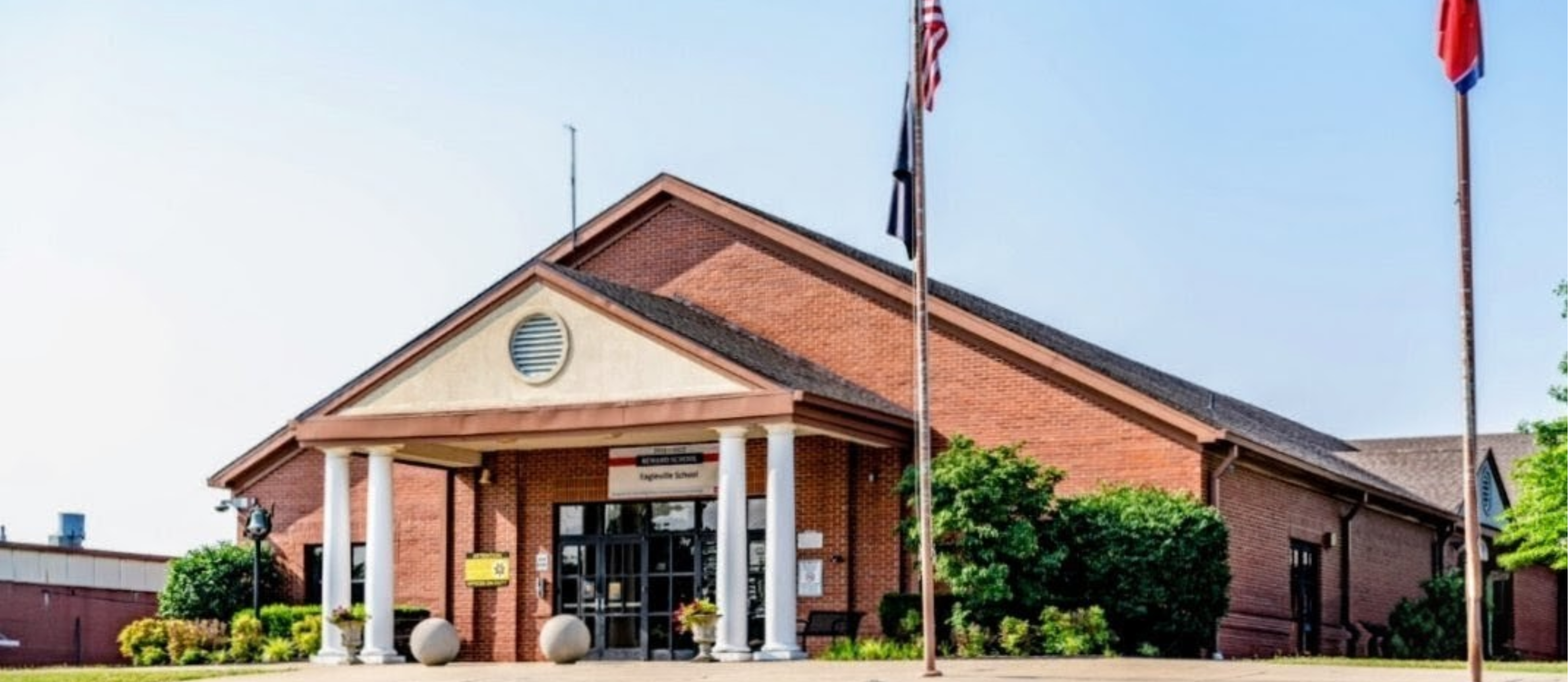 A brick building with a front porch, two flags, and a round window. Plants and trees in front.