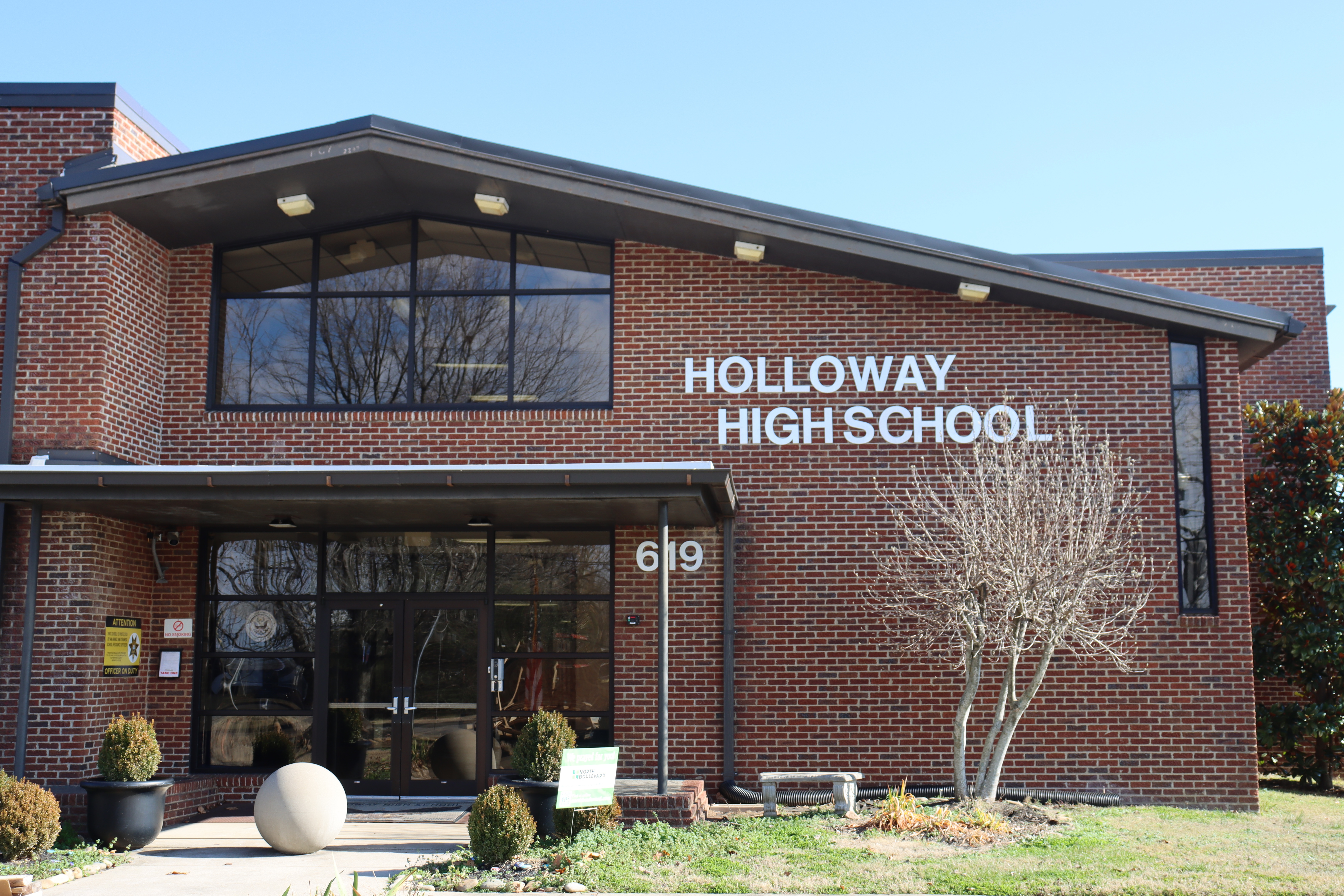 A brick building named Holloway High School with glass doors, windows, a sloping roof, and small trees and plants outside.
