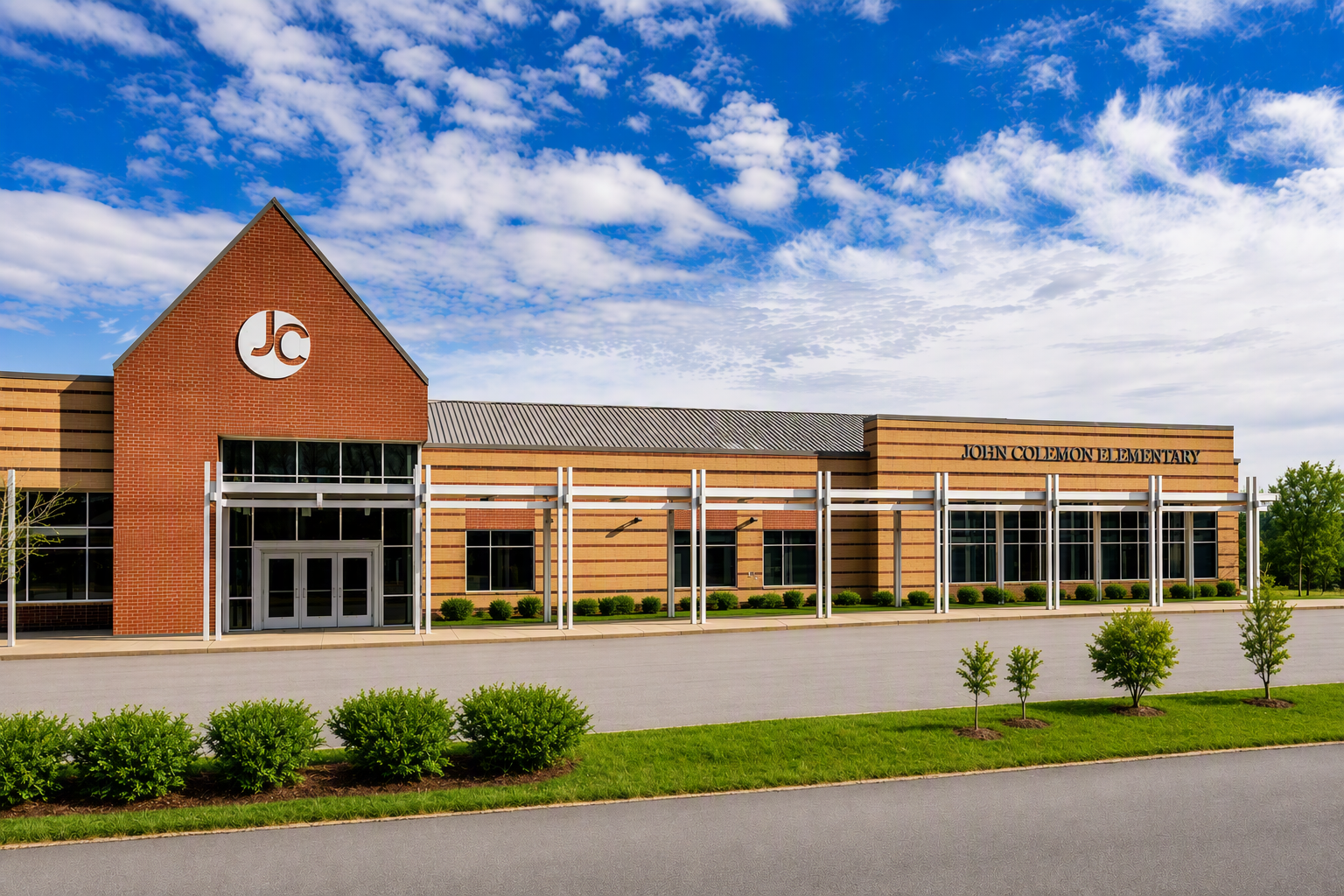 A school building with a logo on the front, glass doors, and a large window. Surrounded by bushes and a green lawn.