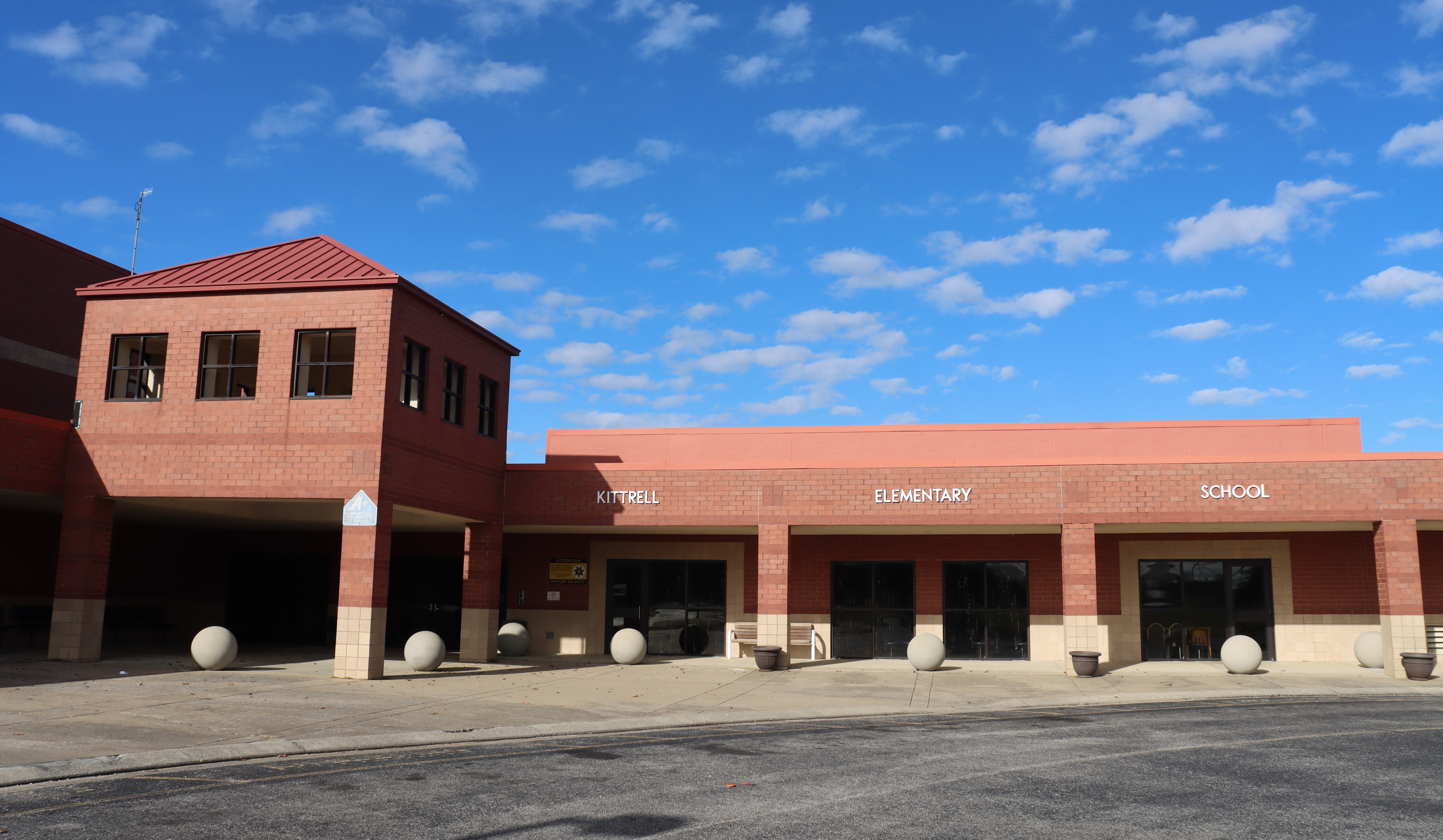 A building with a red roof and brick exterior, with a parking lot in front. Clear blue sky with clouds.