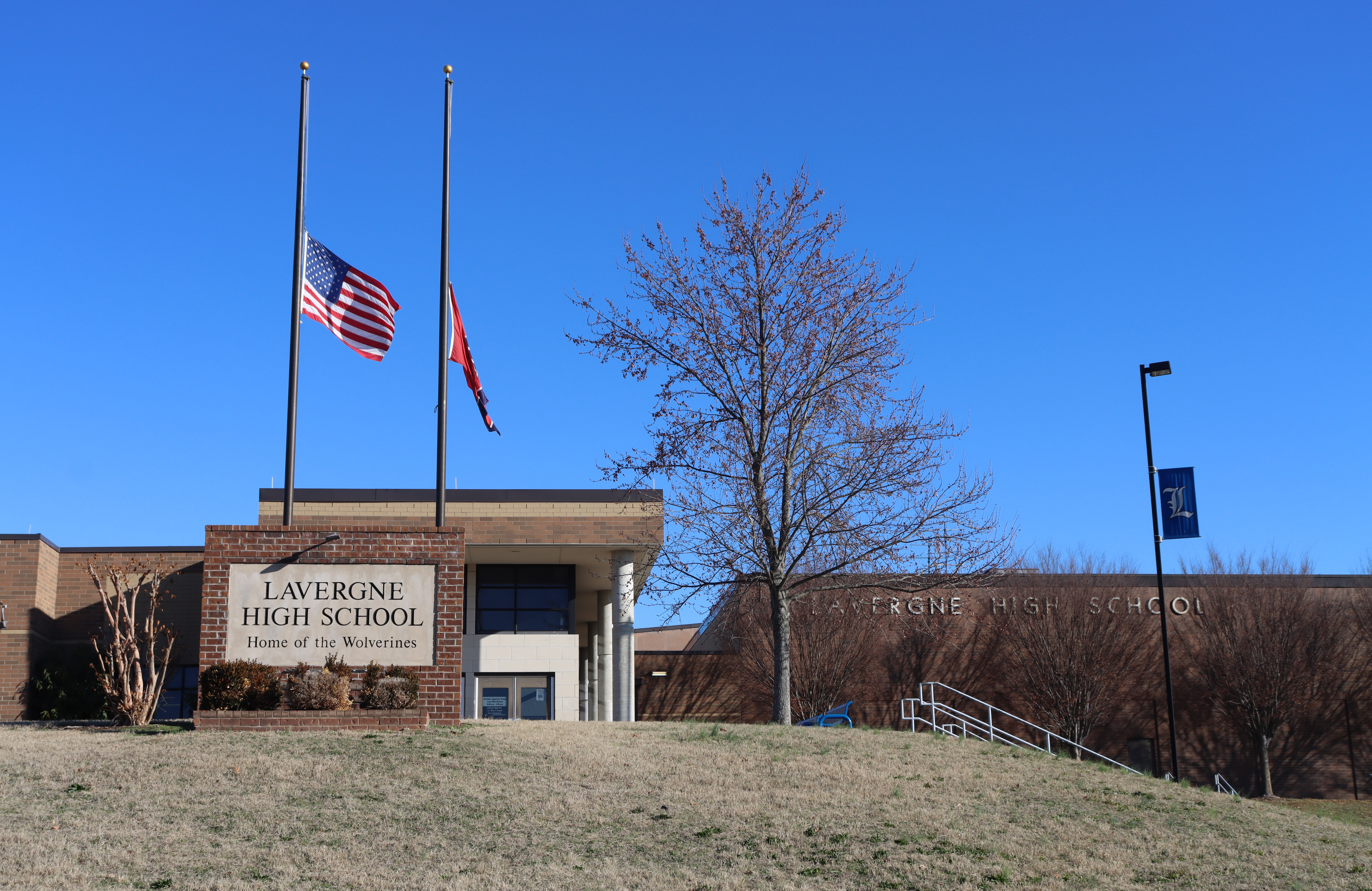 Building entrance with flags on poles, one of which is the American flag. A tree with no leaves.
