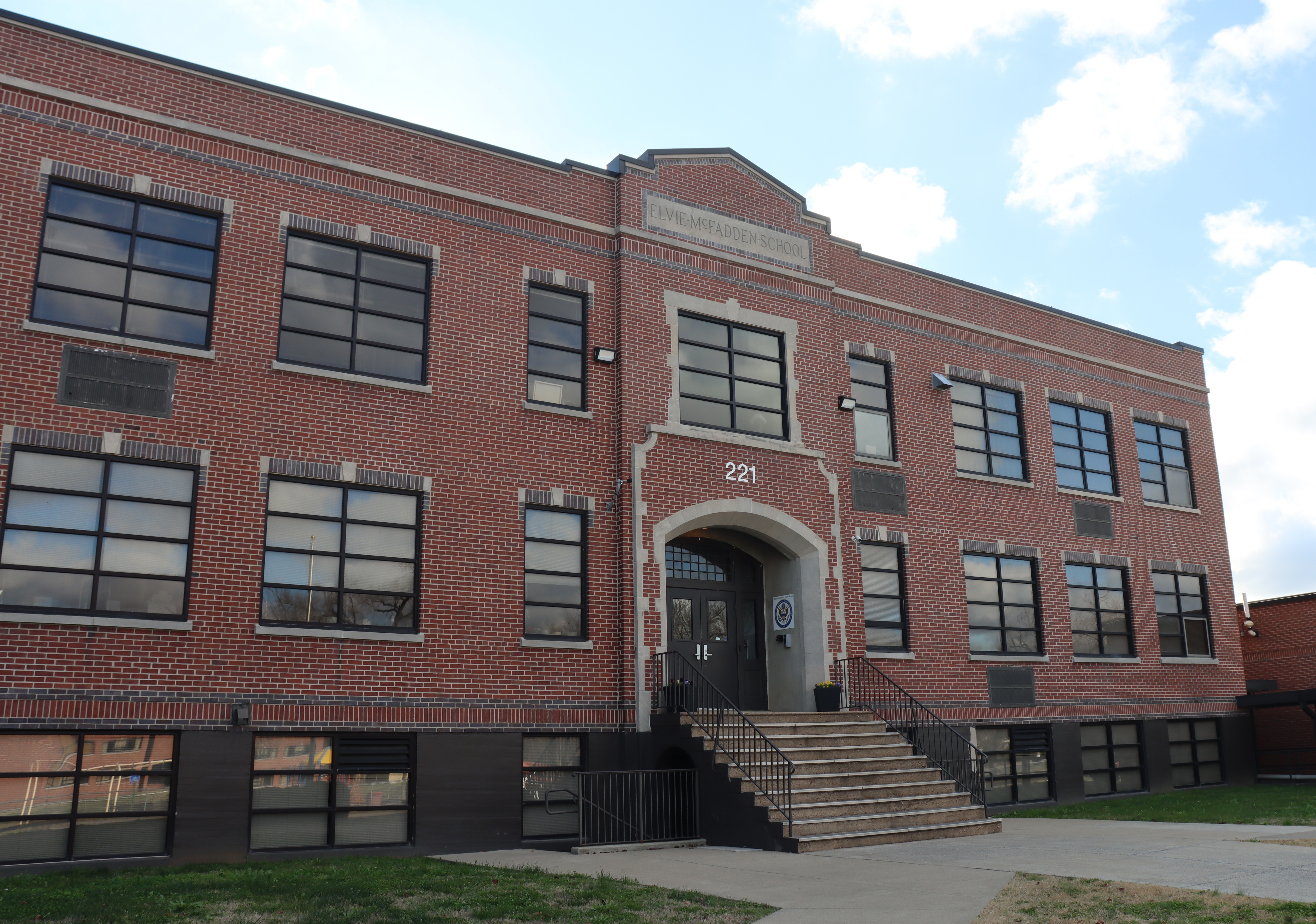 A large brick building with many windows and a wide staircase leading to the entrance, set against a blue sky with clouds.