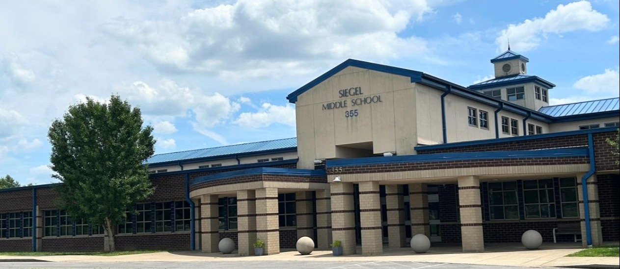 A school building with blue trim and white walls has a blue roof. A tree stands before it.