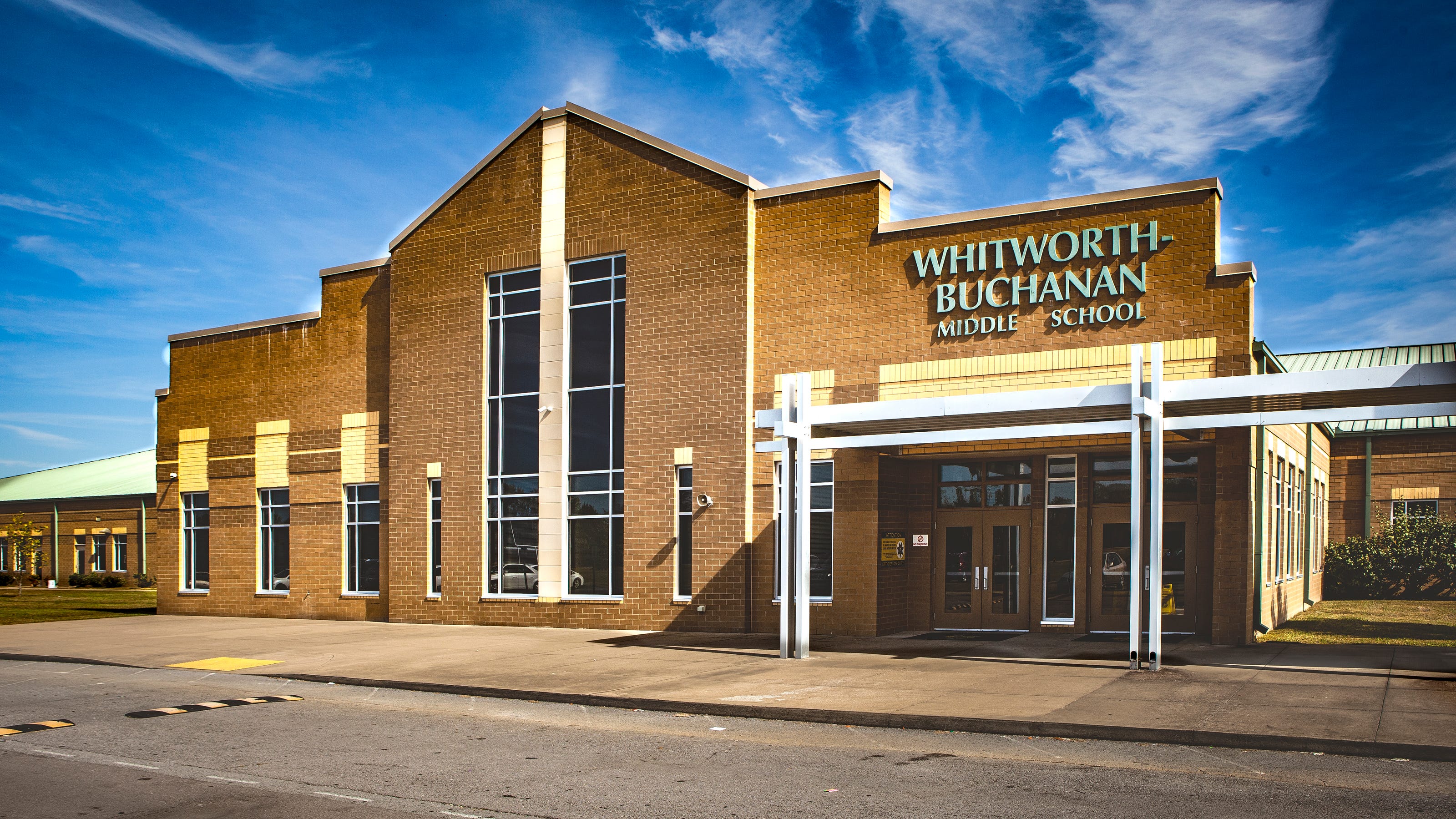 A modern school building with a brown facade, glass windows, and a white sign reading "Whitworth Buchanan School".