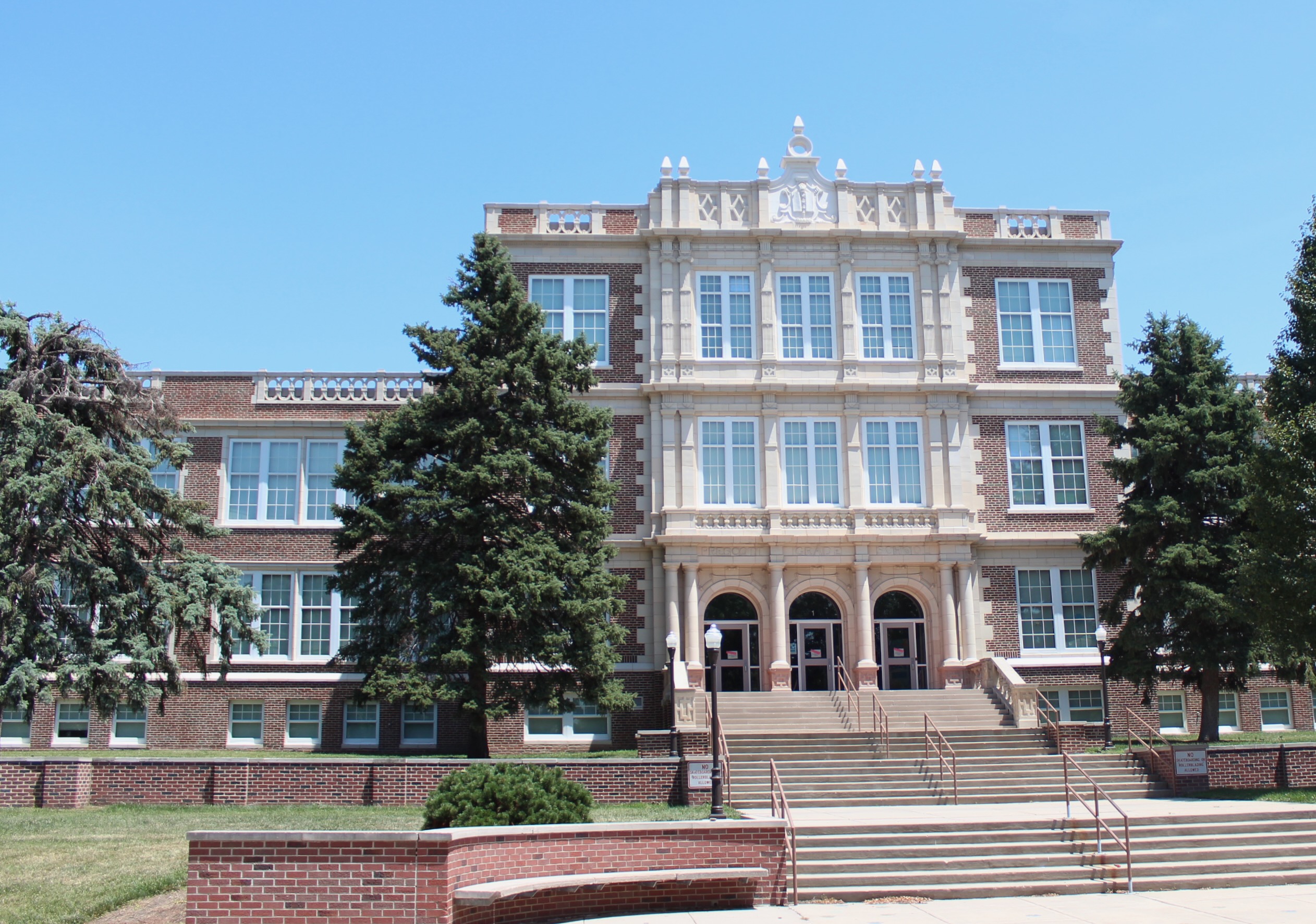Photo of the exterior of Prescott Elementary, a grand brick building with a 3 story center flanked by 2-story wings, against a blue sky
