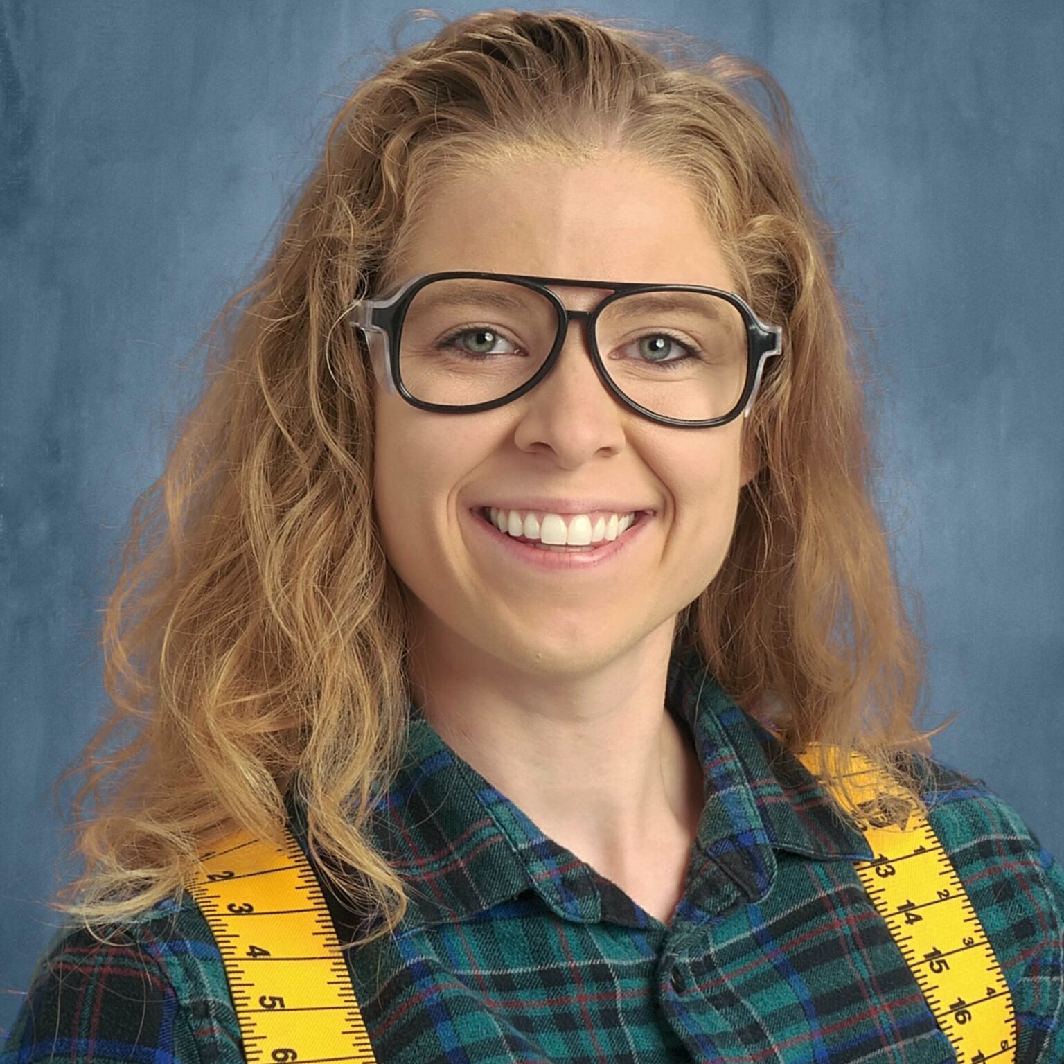 Portrait of woman in work glasses, a green collared shirt and yellow suspenders that look like rulers. 
