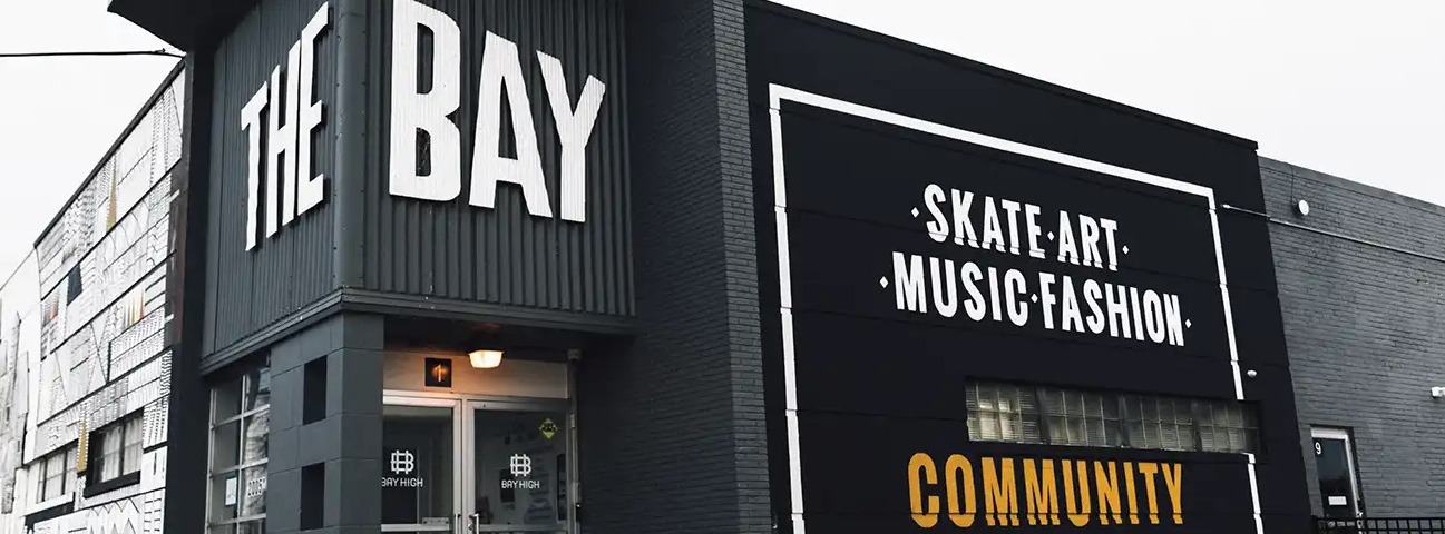 The exterior of a dark gray industrial building with large white lettering reading "The Bay" above a glass double-door entrance. A painted sign on the side wall reads "Skate, art, music, fashion" with "Community" in gold below it, and a black-and-white mural covers the far end of the building under an overcast sky.
