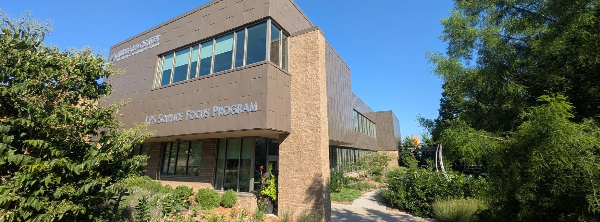 A two story building, colored in earth tones of light and medium brown nestled in trees on each side. Signage on the building says "LPS Science Focus Program"