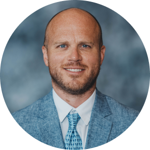 Photo of bald man with light facial hair in a blue jacket, white shirt and blue tie over a blue-gray background