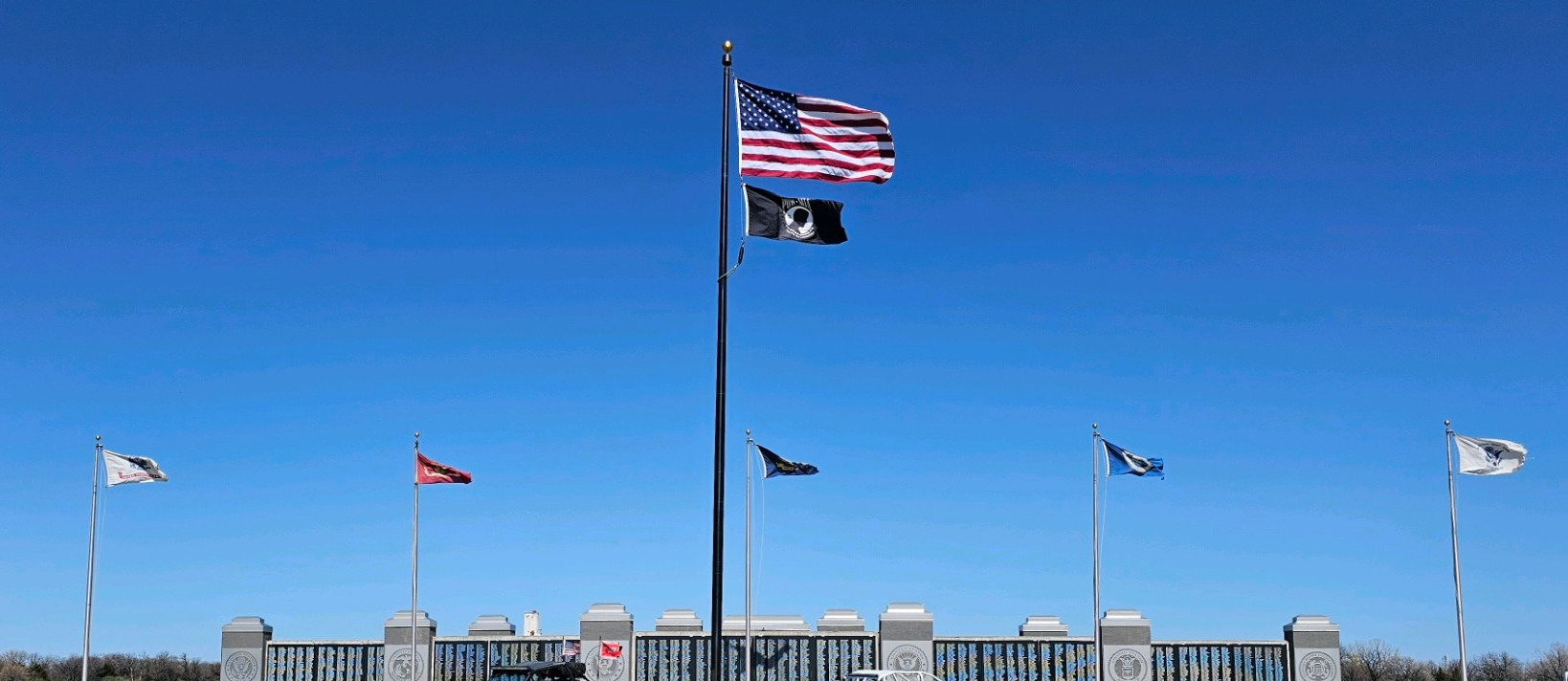 American and POW/MIA flags fly on a tall central flagpole in front of a military memorial wall engraved with names, with additional service flags on either side and two off-road utility vehicles parked on the brick plaza under a clear blue sky.