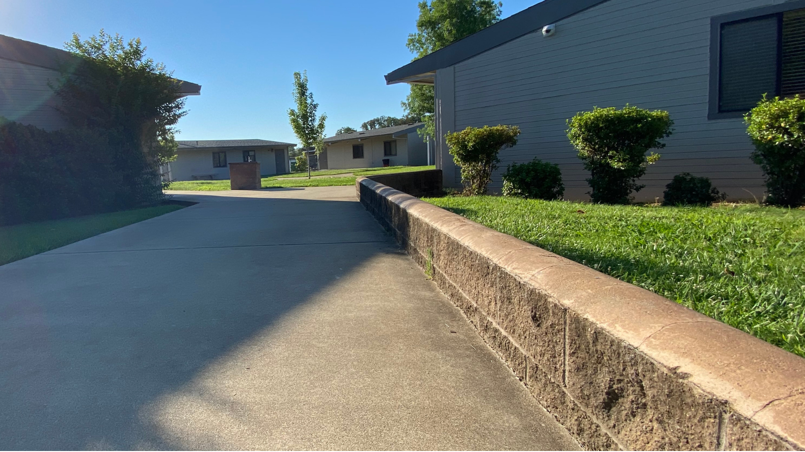 Concrete driveway with a grassy median and a small trash bin. Buildings and greenery line the background.