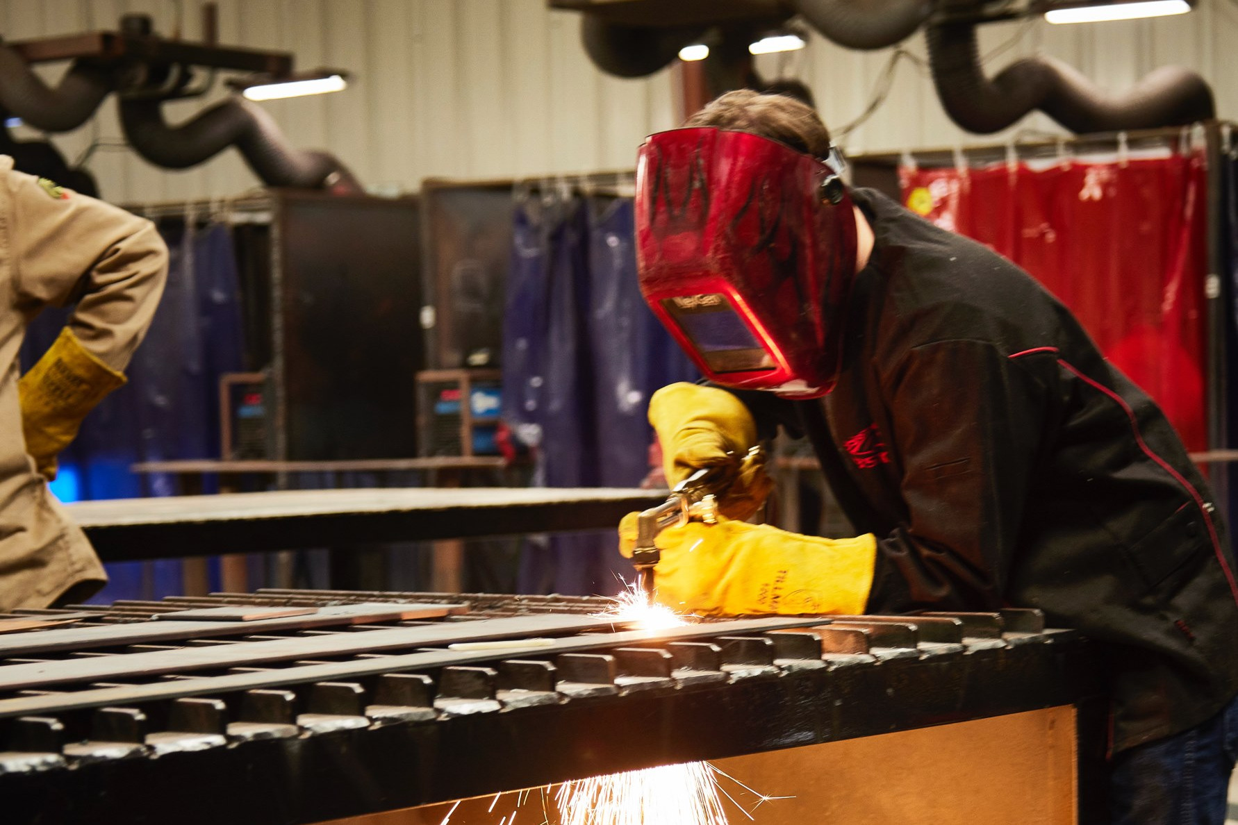 Person in welding mask and gloves cuts metal on a bench. Sparks fly. Industrial setting.