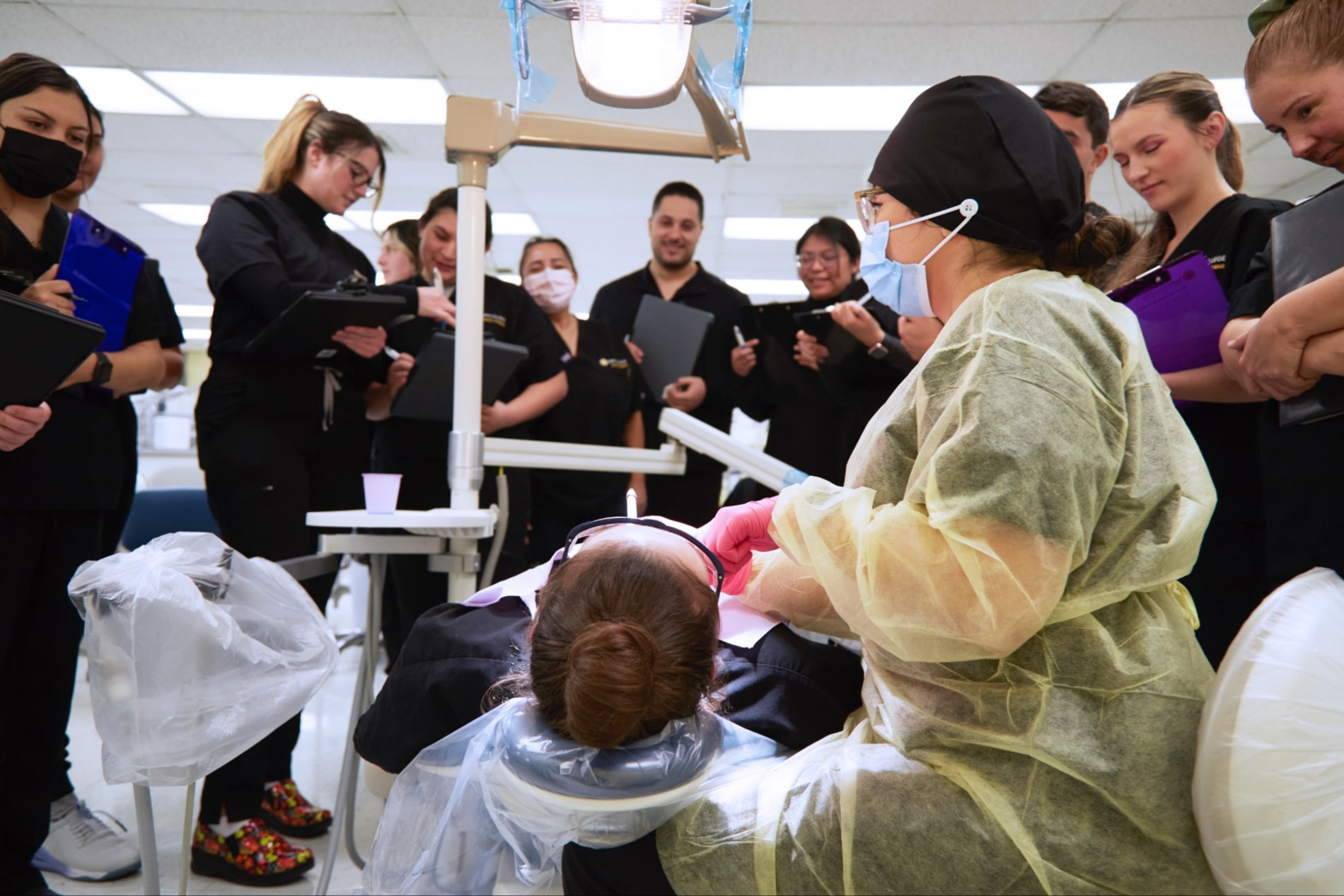 A dentist in protective gear examines a patient seated in a dental chair. Others observe, some masked.