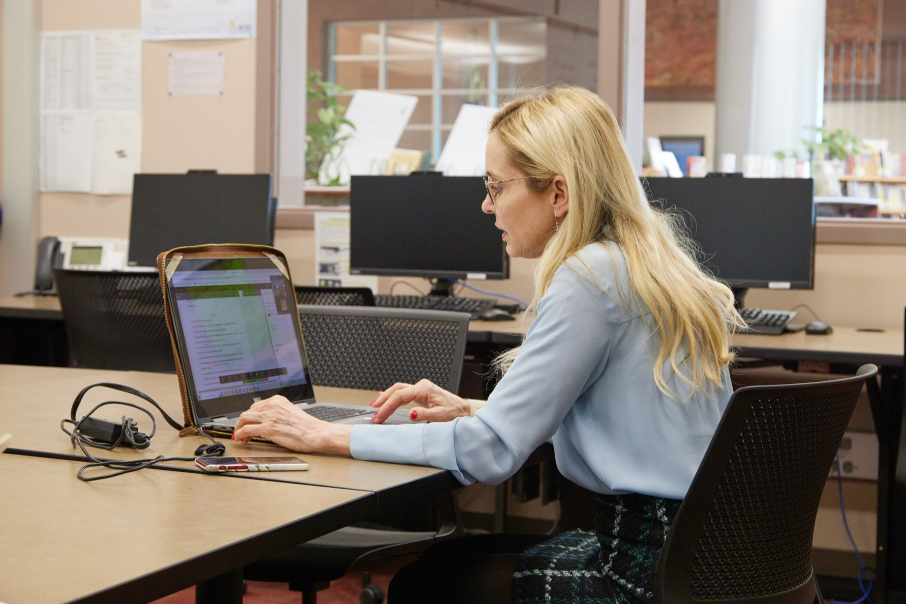 Woman in a long-sleeve shirt and skirt seated at a desk, typing on a laptop. Behind her, computer monitors are visible.