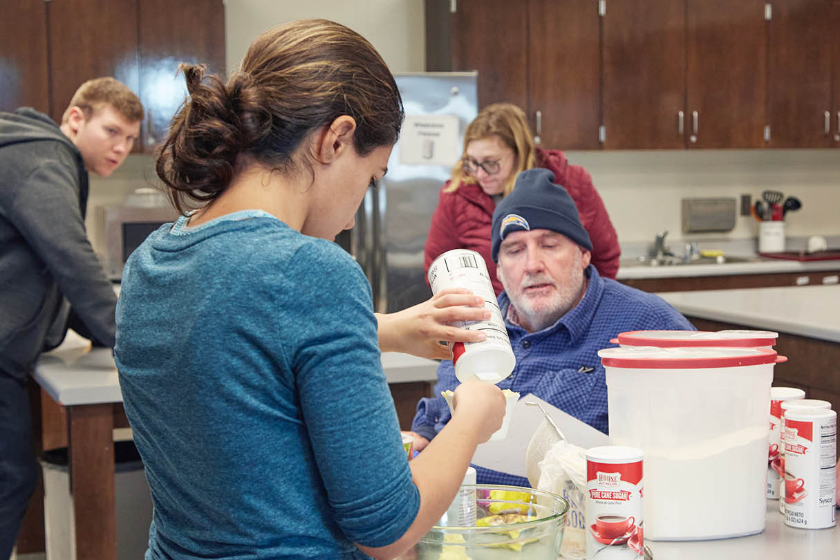 People prepare food in a kitchen. One person pours a liquid into a plastic container.