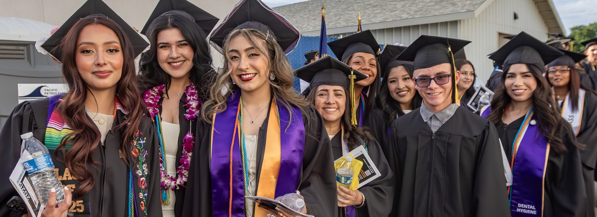 A group of people in caps and gowns pose for a photo outdoors with buildings in the background.