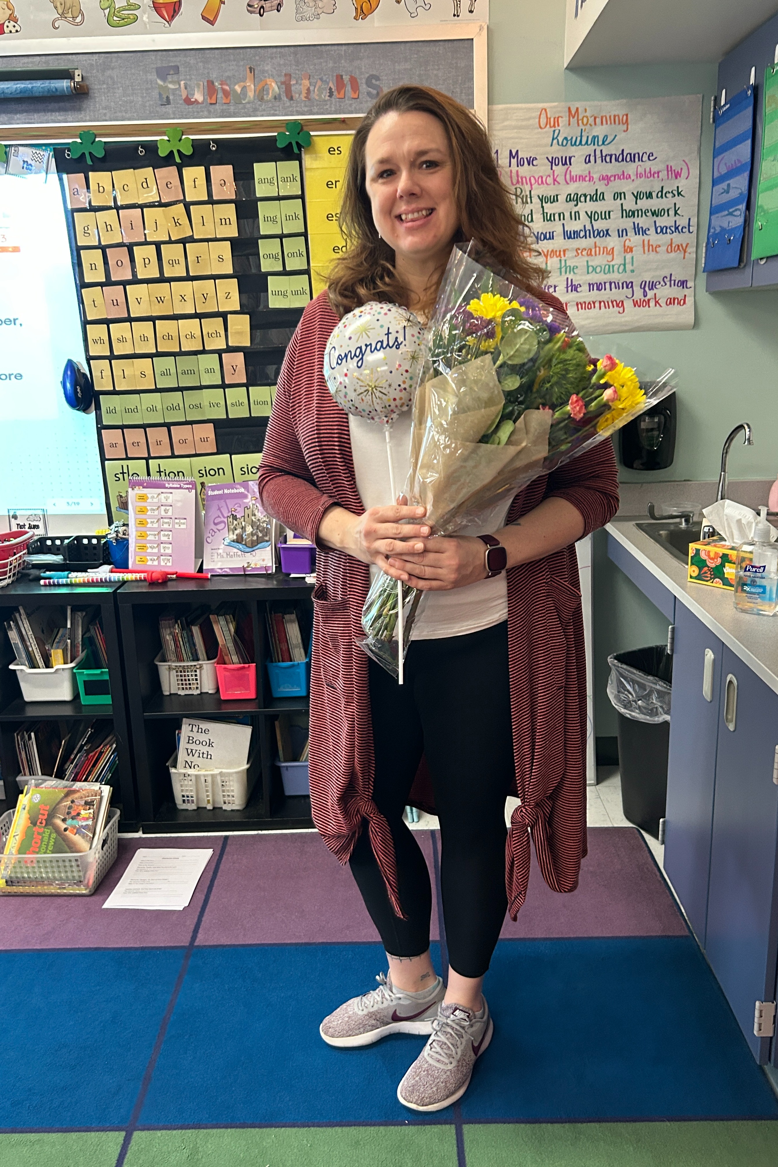 A woman holds a bouquet of flowers in a classroom with a bookshelf, a sink, and a whiteboard.