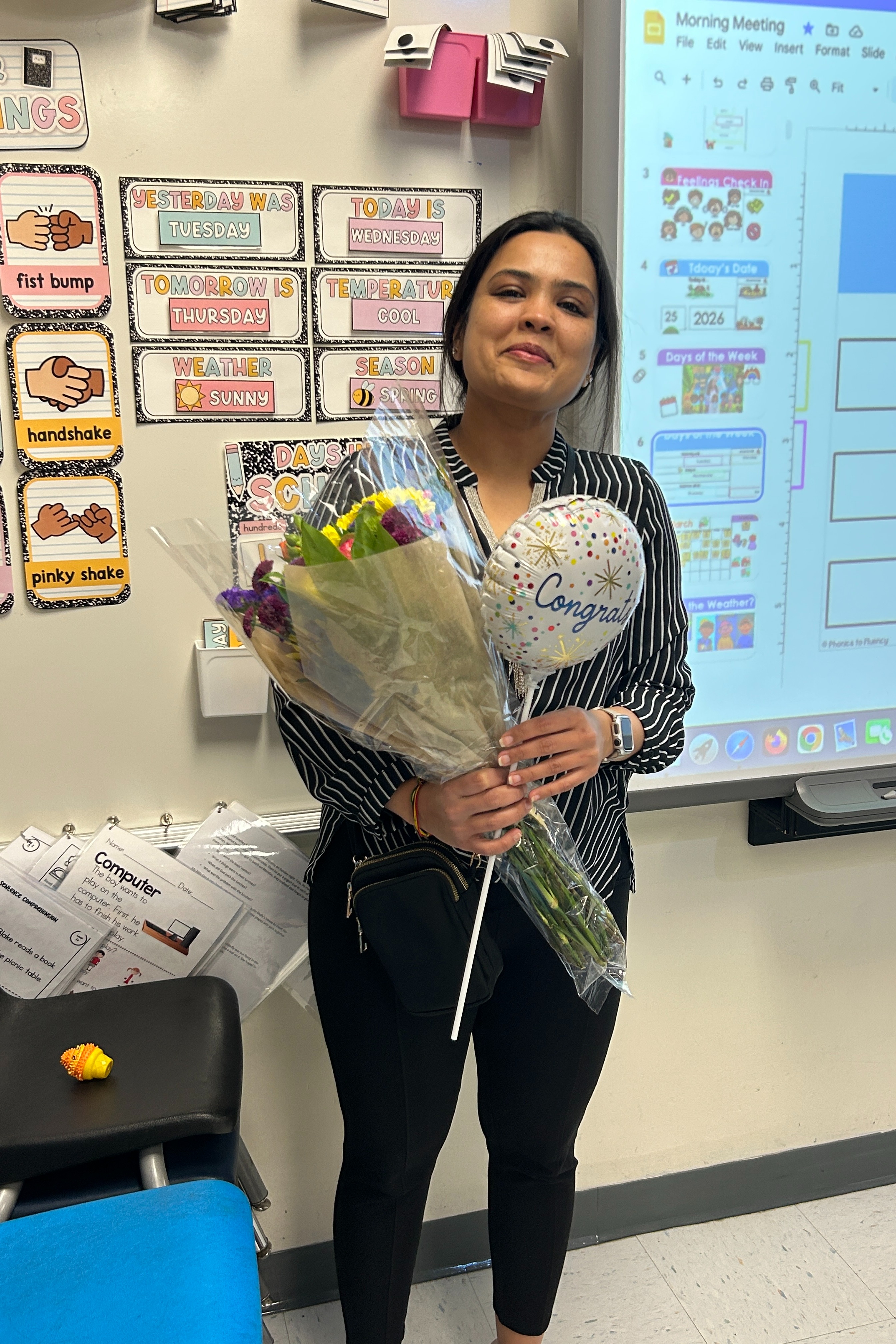 A woman stands in a classroom holding a bouquet of flowers and a balloon, with a projector screen behind her.