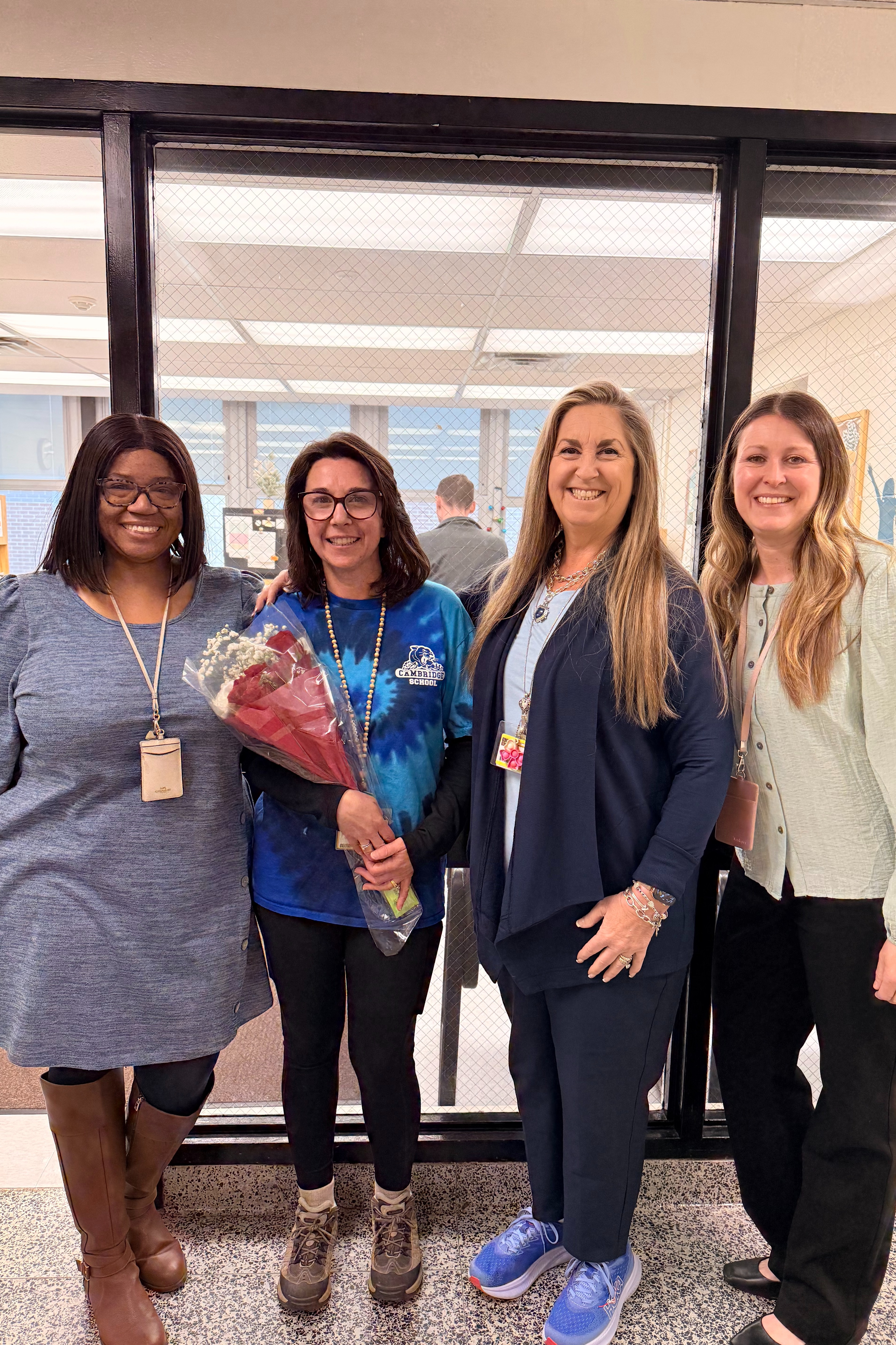 Four women stand in front of a glass door. The woman on the left holds a bouquet of flowers.
