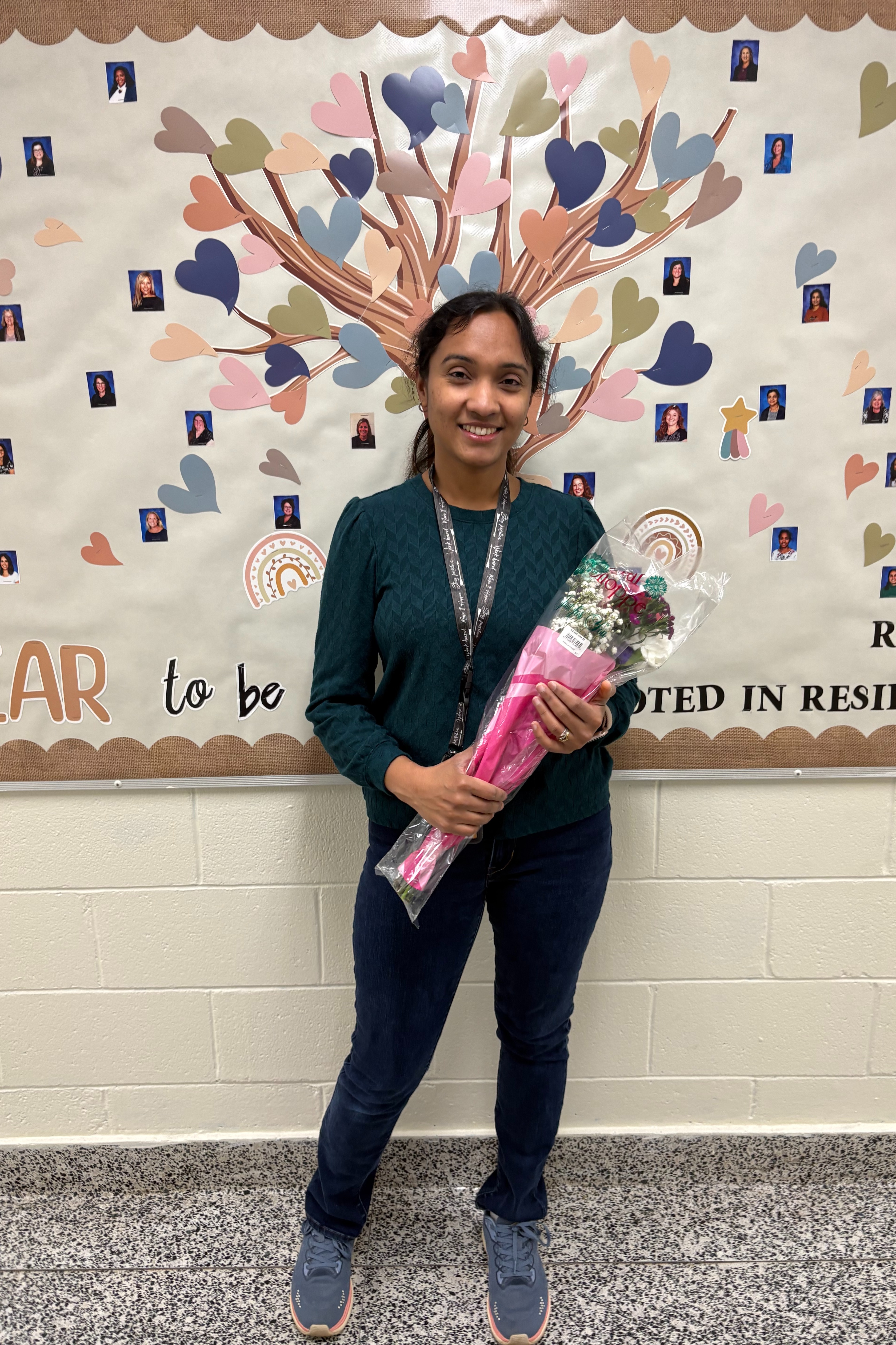 Person with long dark hair standing in front of a decorated wall, holding a bouquet of flowers.
