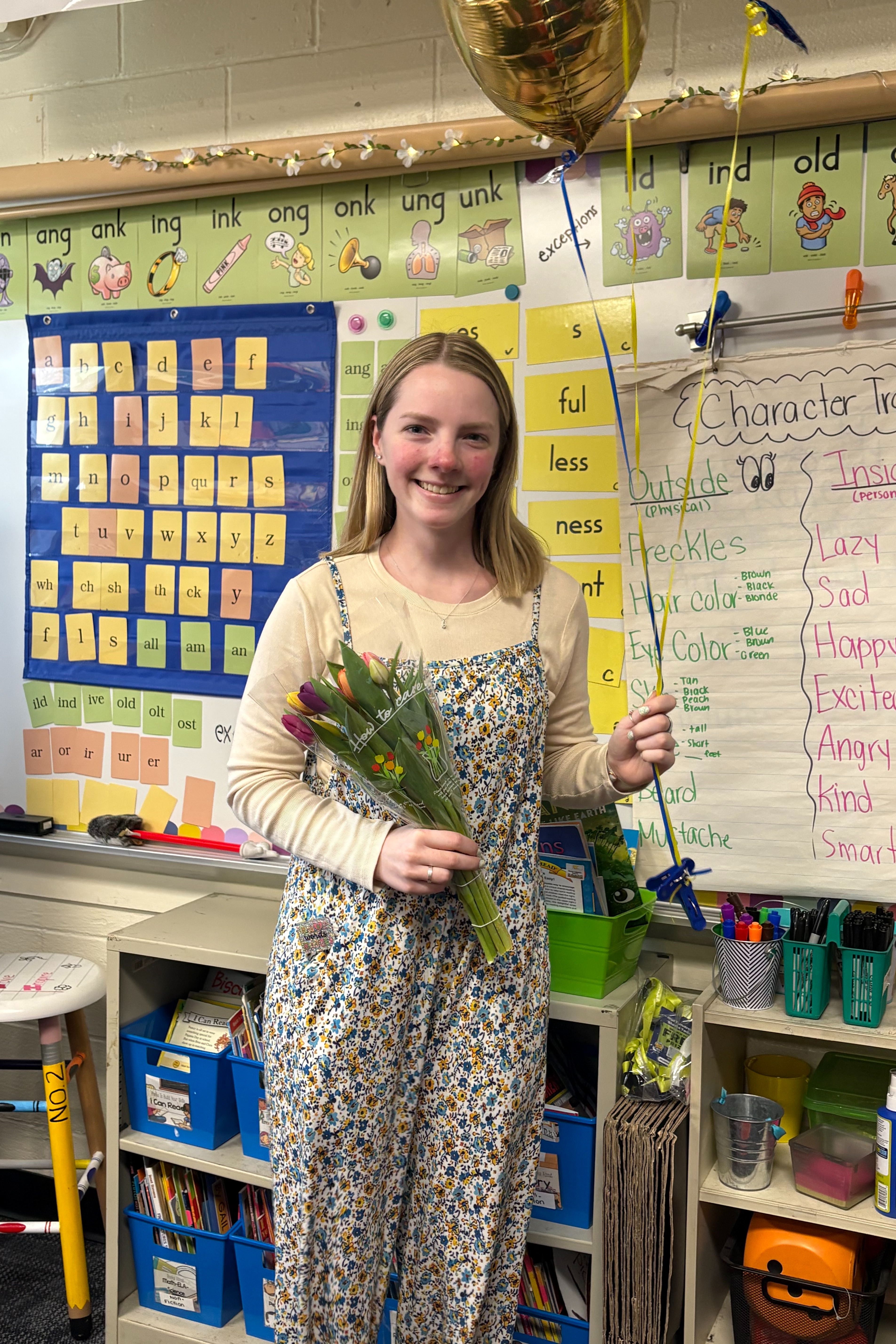 A woman in a floral jumpsuit holding a bouquet of flowers stands in a classroom.