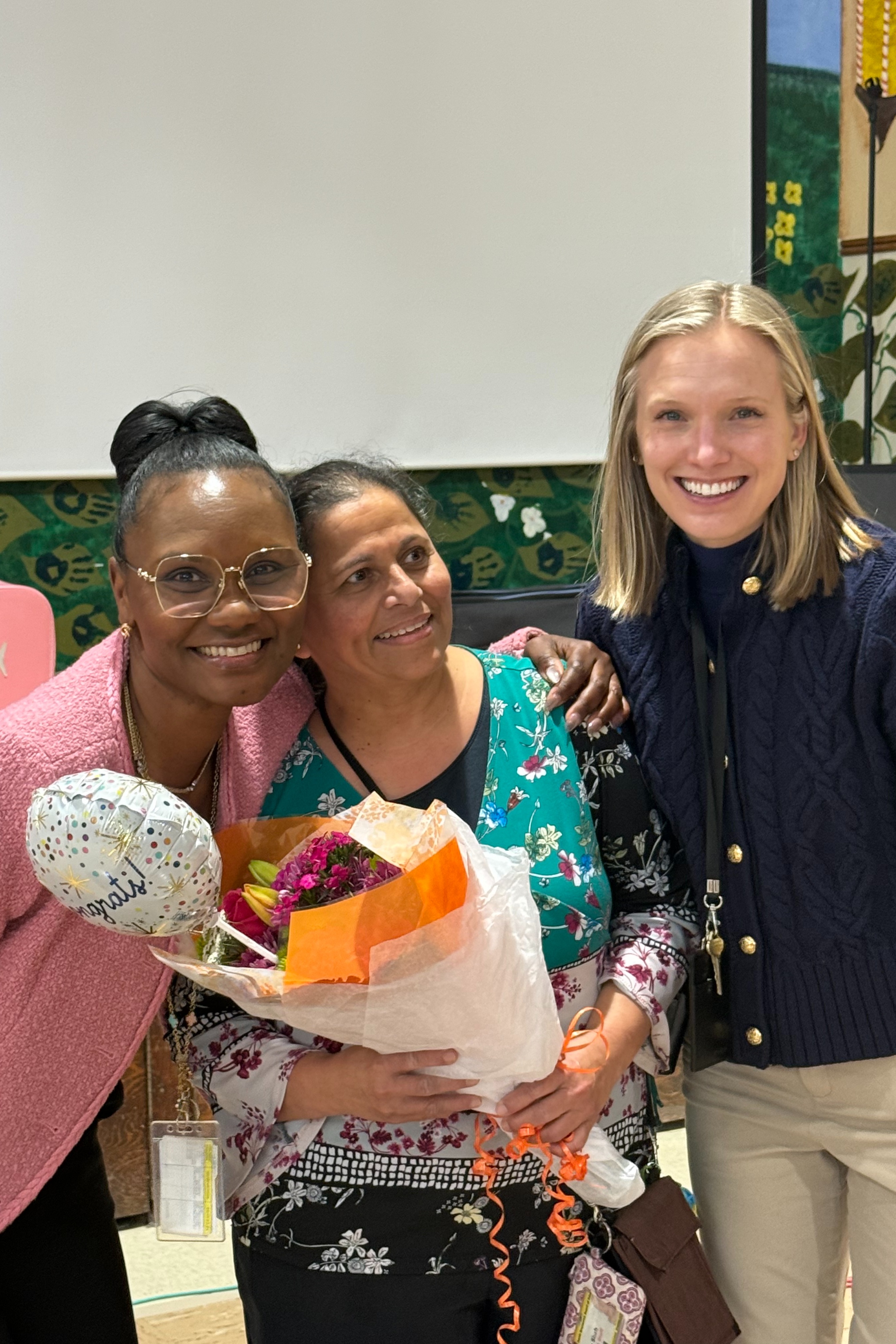 Three women stand together, one holding a bouquet of flowers, with a mural in the background.