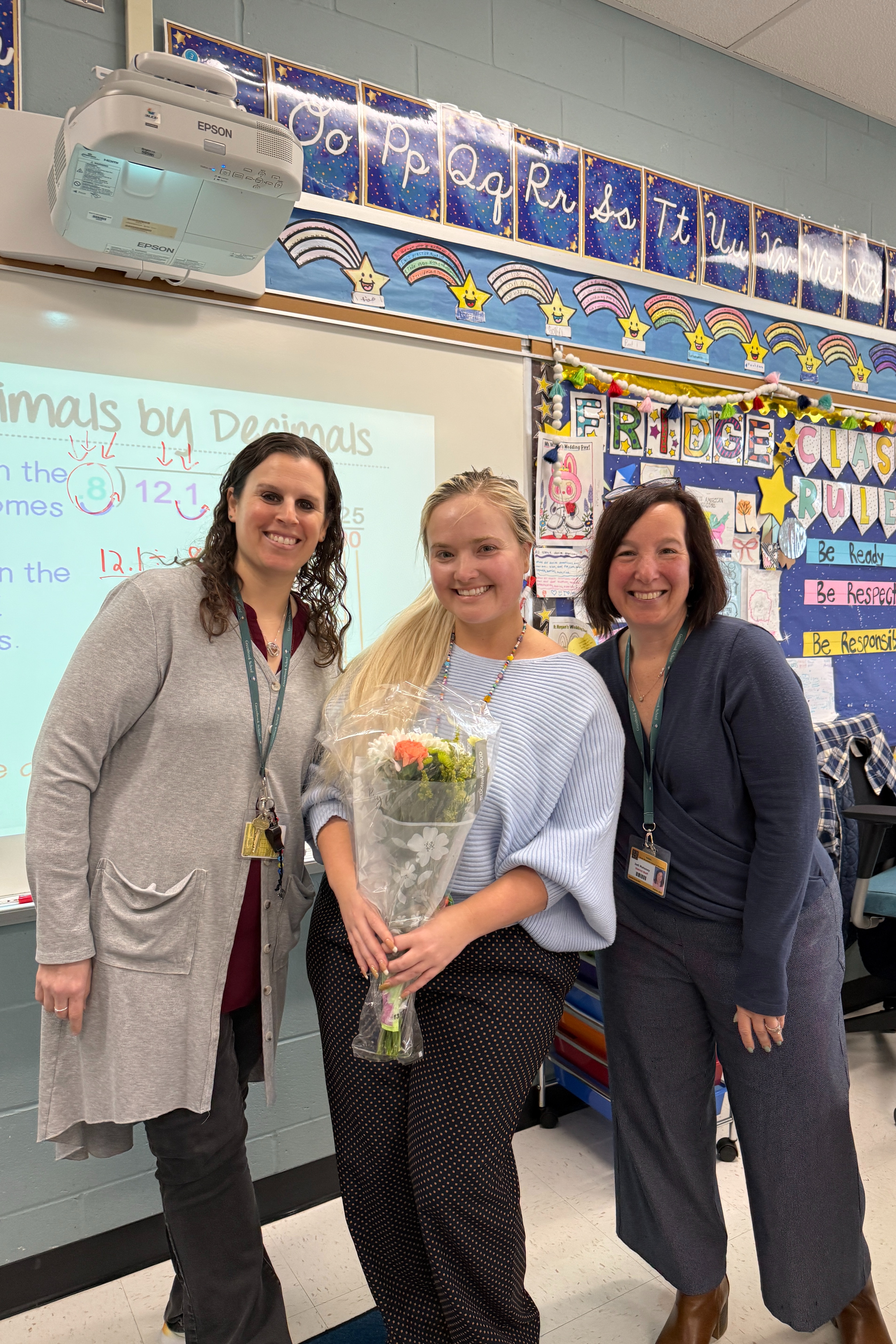 Three women stand in a classroom. The middle woman holds flowers. A whiteboard is behind them with colorful decorations.