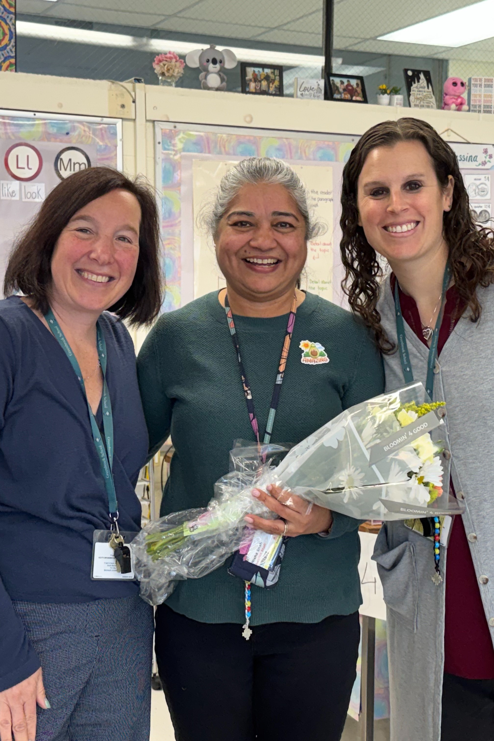Three women stand in a classroom, smiling. The woman on the right holds a bouquet of flowers.