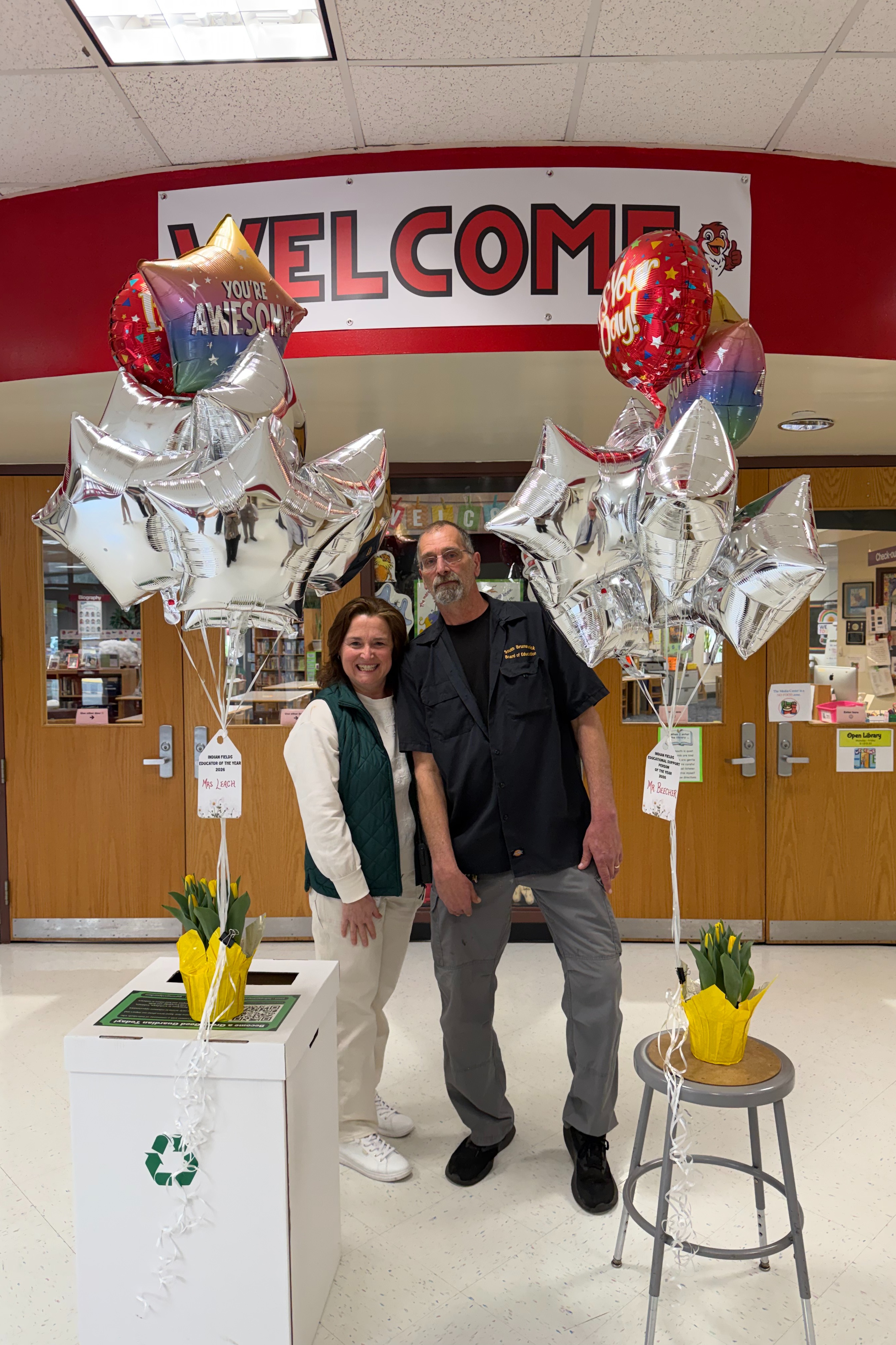 A man and a woman stand next to a recycling bin and a stool with balloons in front of a building.