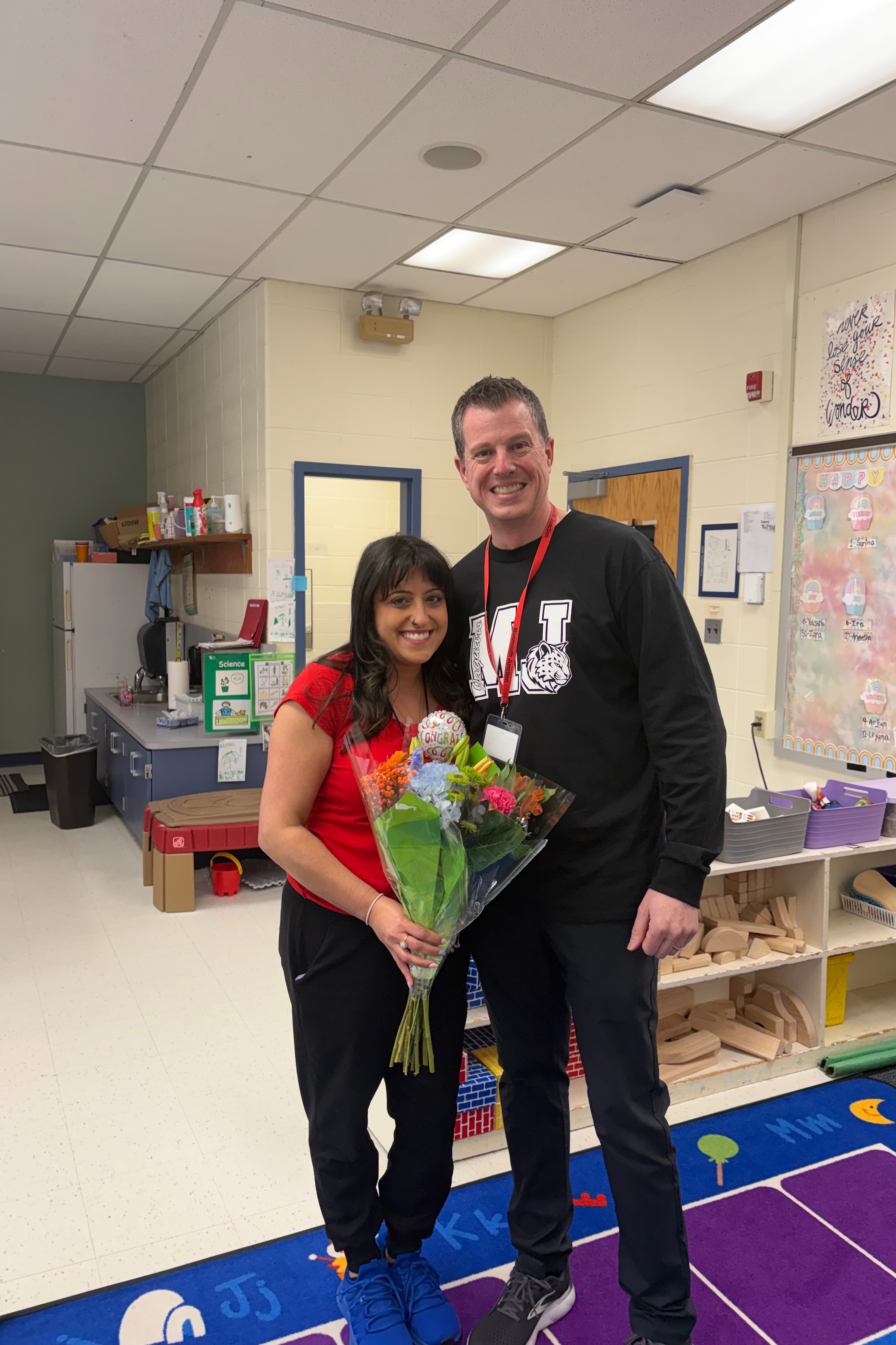 A man and woman stand together in a room. The man holds a bouquet of flowers. The floor is covered with a rug.