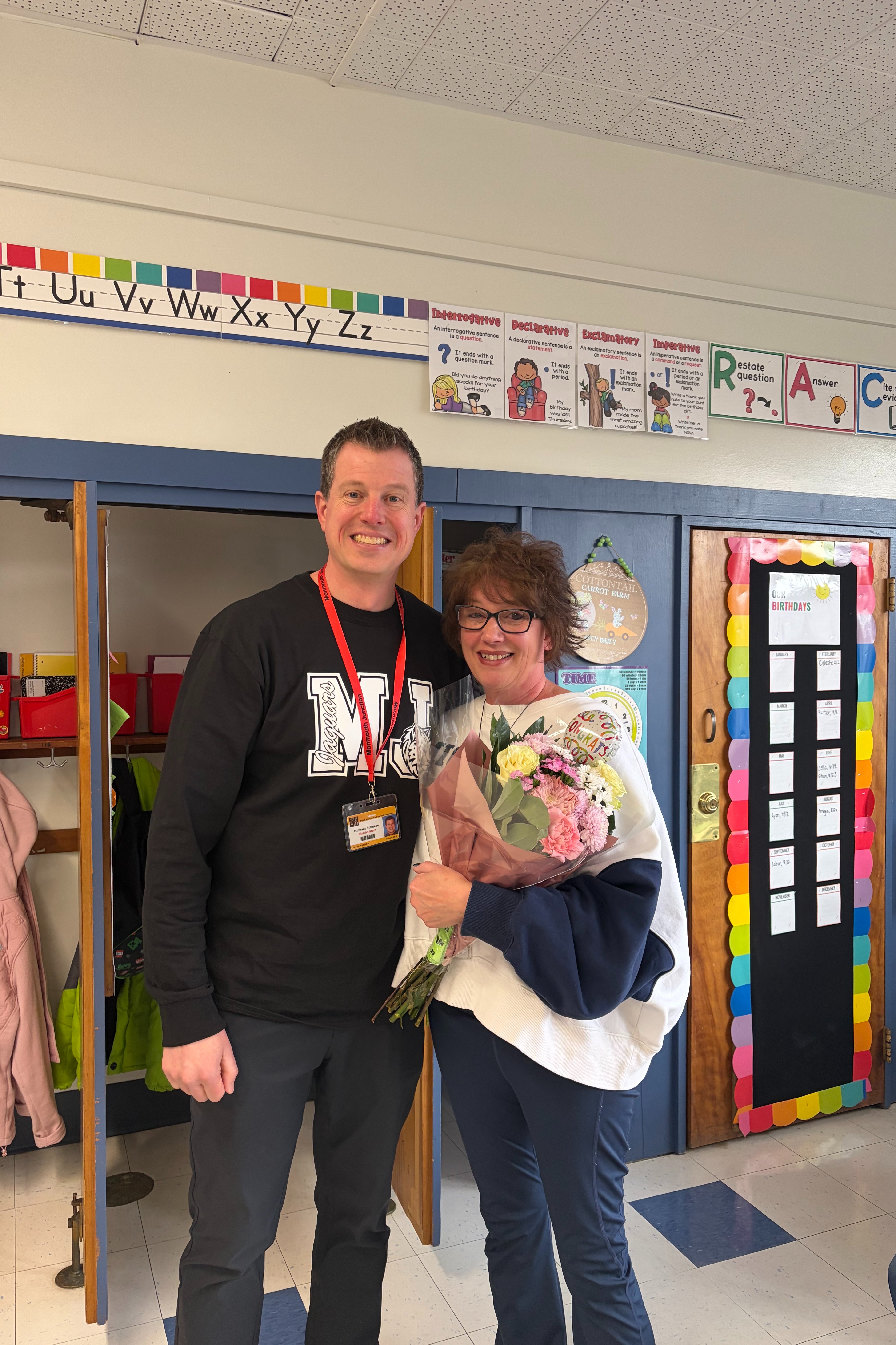 A man and woman stand in a classroom. The woman holds flowers, and they both smile. Behind them are colorful classroom decorations.