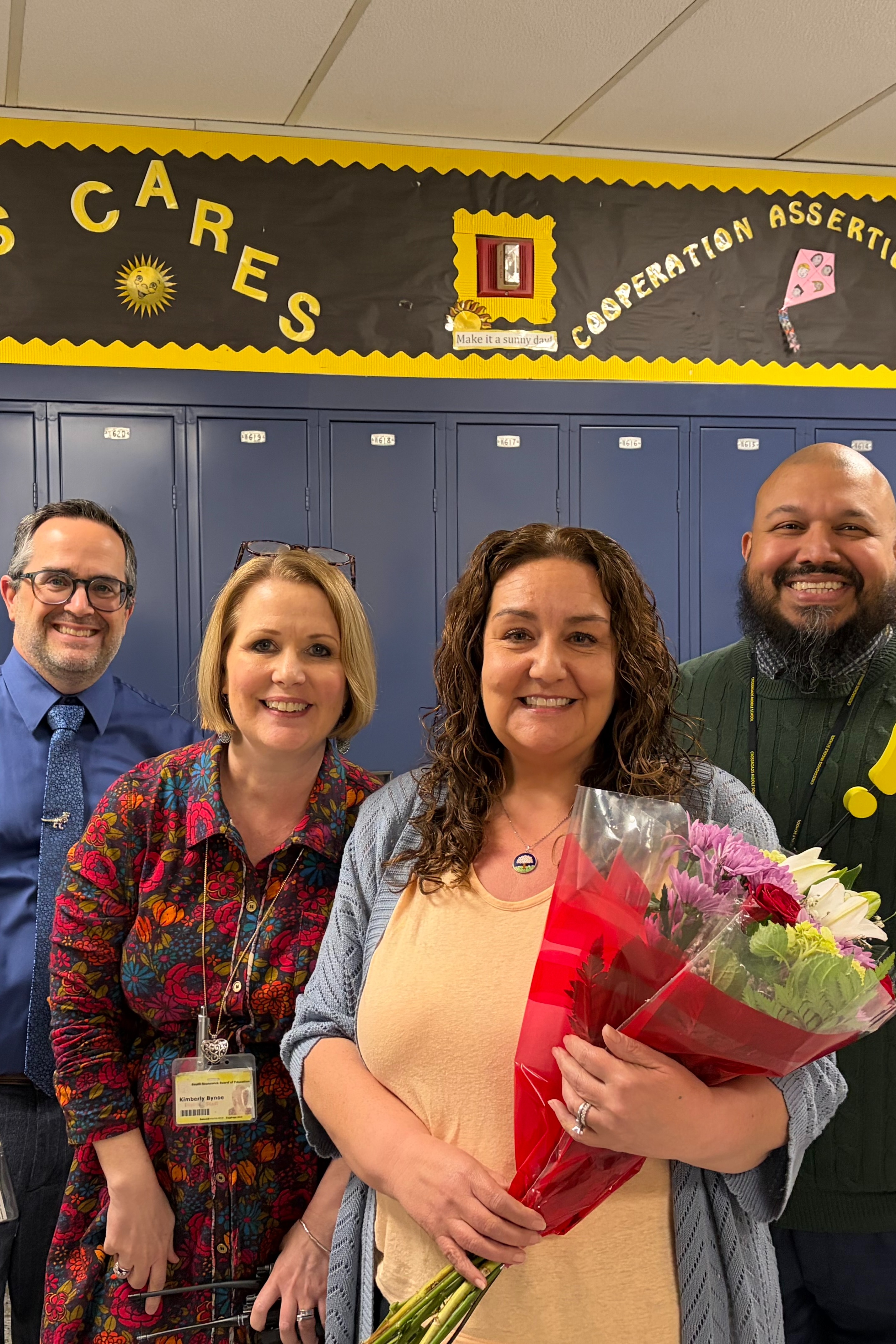 Five people in a school hallway, with one person holding a bouquet of flowers. Behind them, lockers line the wall with a sign reading "CROSSROADS CARES."
