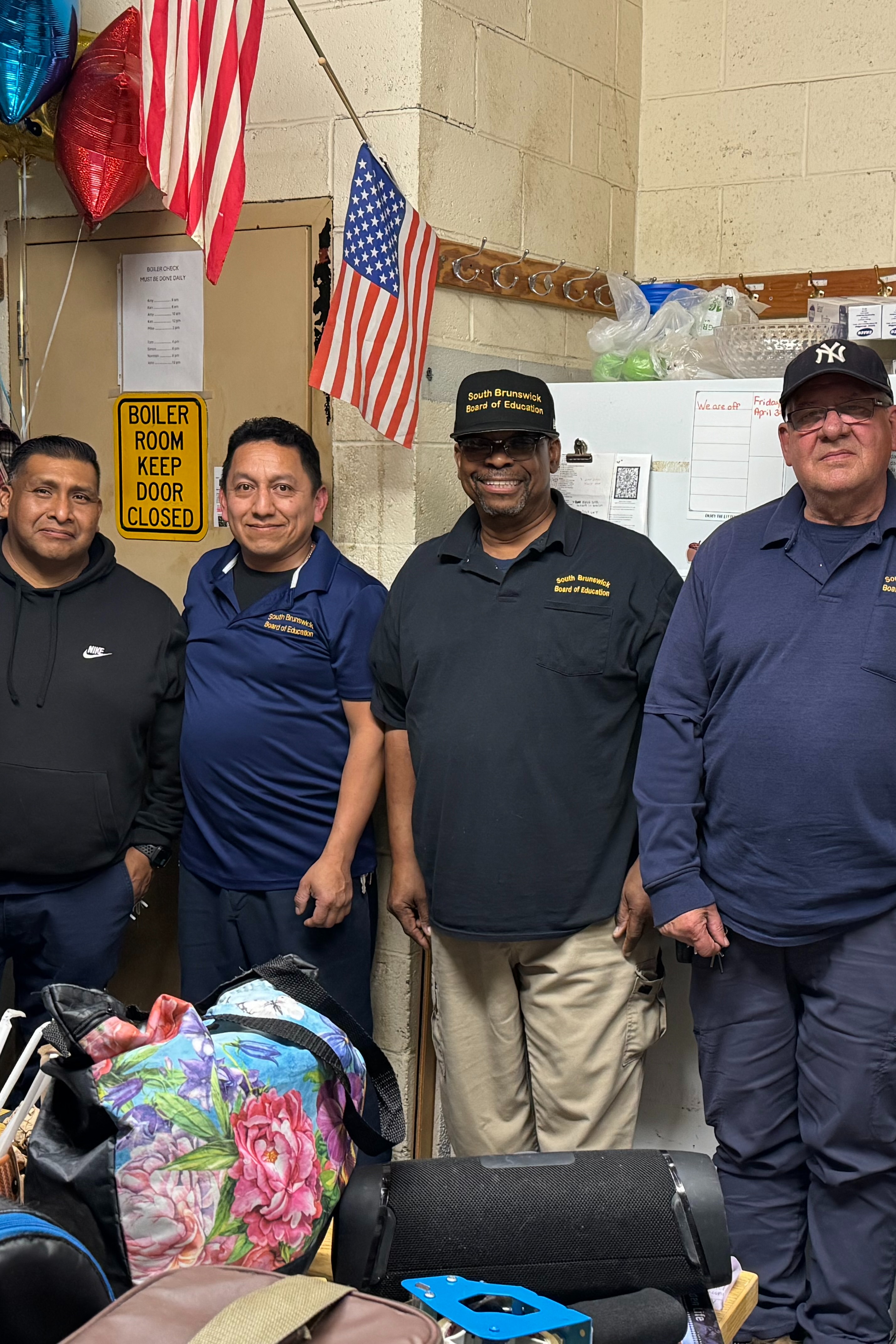 Six men in uniform stand in a room with shelves, bags, and a flag. They face the camera.