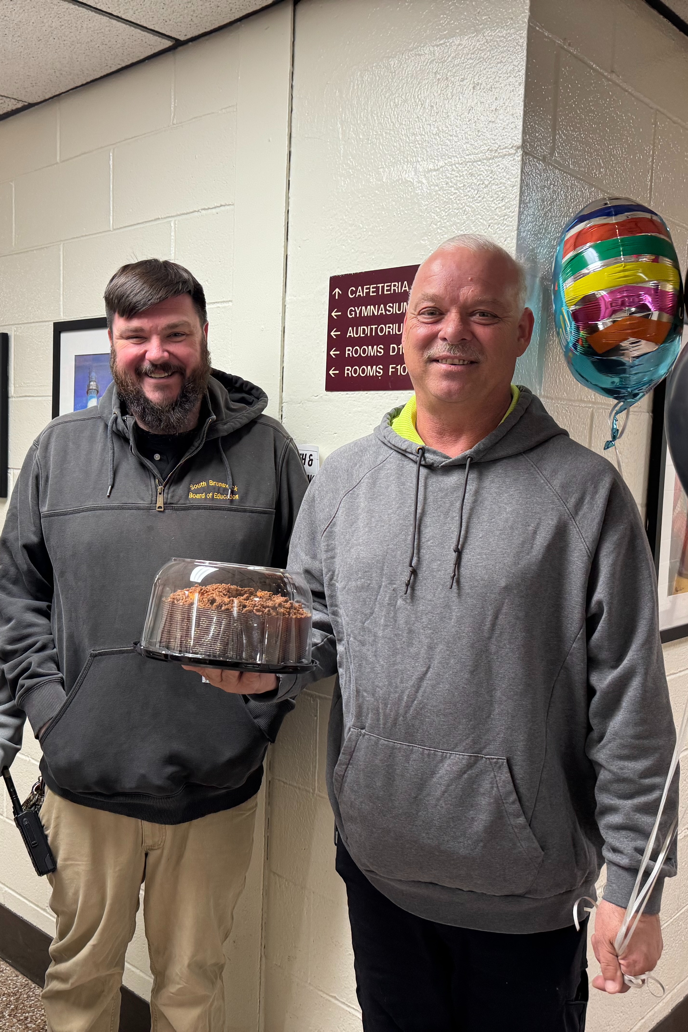 Three adults stand in a corridor, with two men holding a dessert and balloons, while a woman smiles in the background.