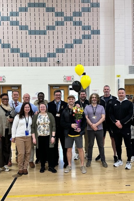 Several people in a gymnasium, some wearing sneakers, one holding a bouquet, balloons behind them.