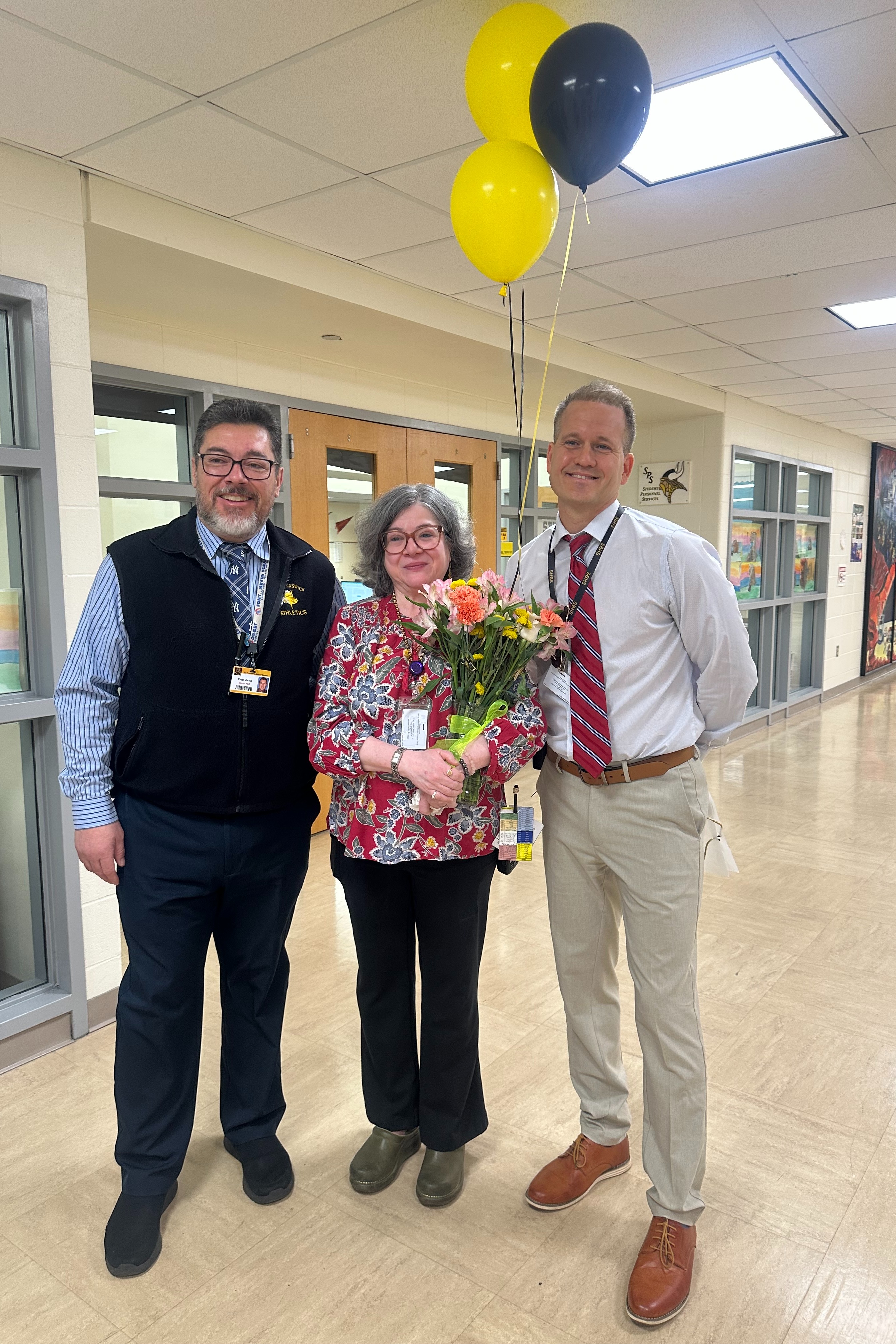 Three people stand in a hallway with yellow balloons. The woman holds a bouquet of flowers.