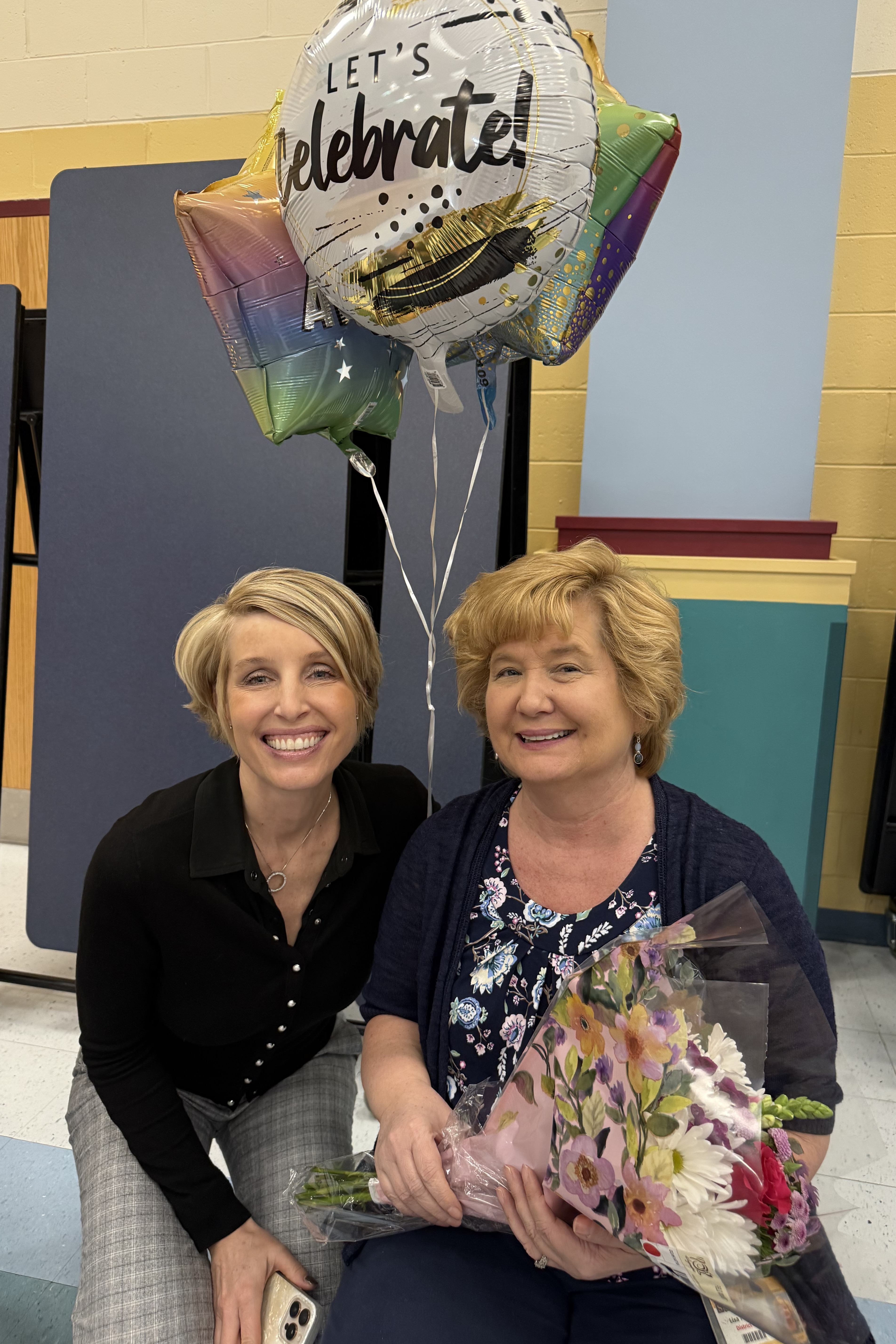 Two women smiling and posing for a photo, one holding a bouquet of flowers, next to a colorful balloon.