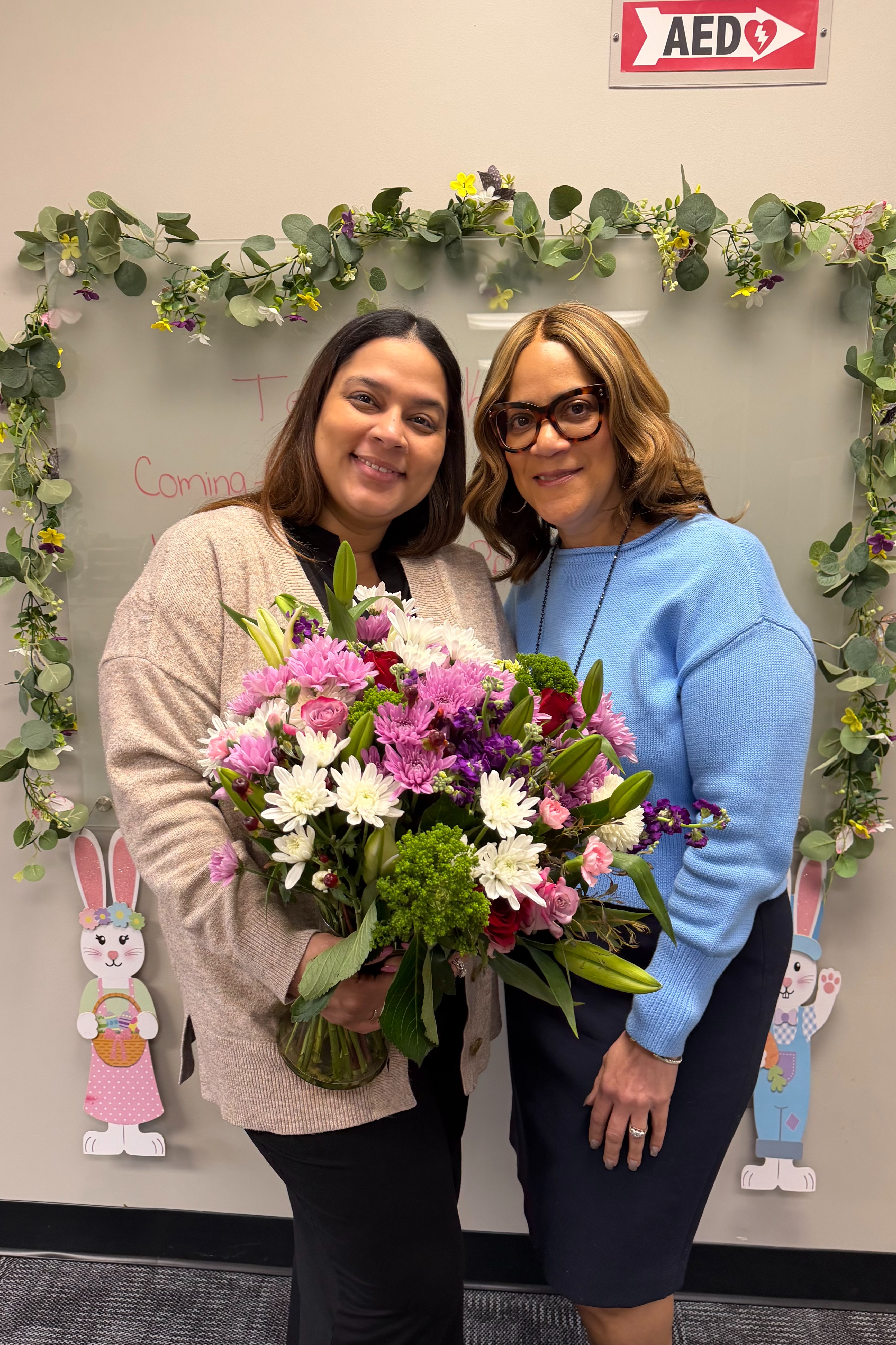 Two women, one holding a bouquet, standing next to each other in front of a heart-shaped flower arrangement.