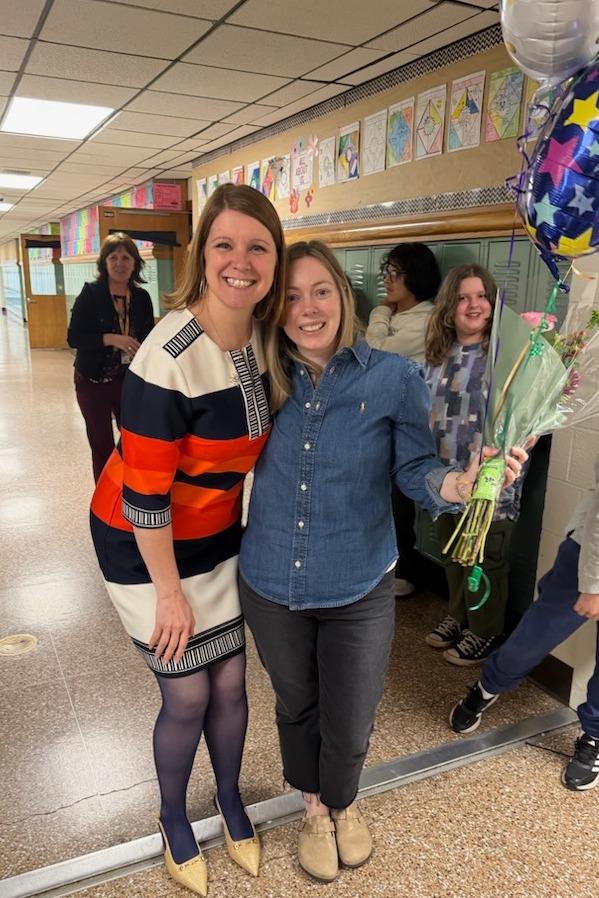 Two women stand together in a hallway, smiling. One holds flowers, the other a balloon. A child stands nearby.