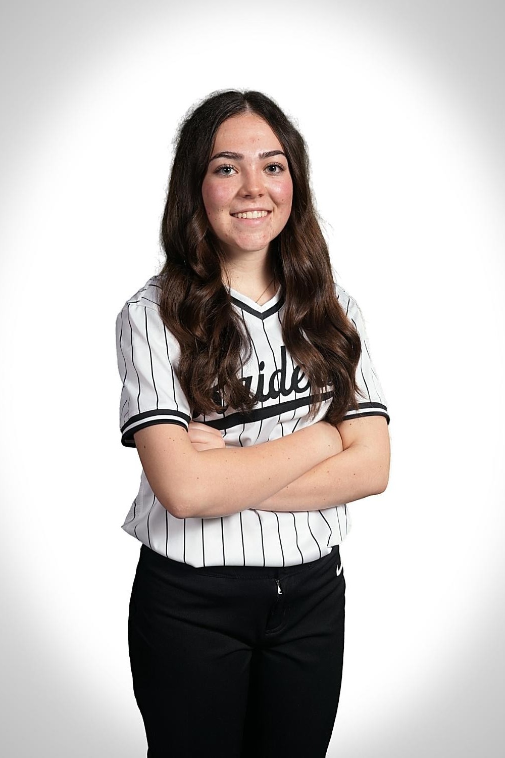 Woman with long hair in a baseball uniform, standing with arms crossed against a plain background.