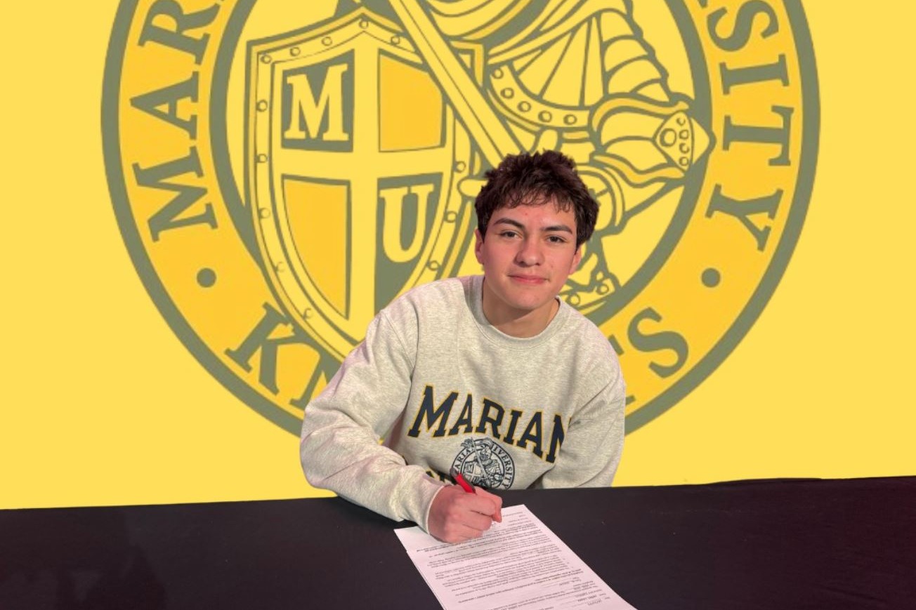 A young man sits at a desk with a document, holding a pen. Behind him, a yellow backdrop displays "SIGNED" and "BOYS SOCCER" with the Marian University logo.