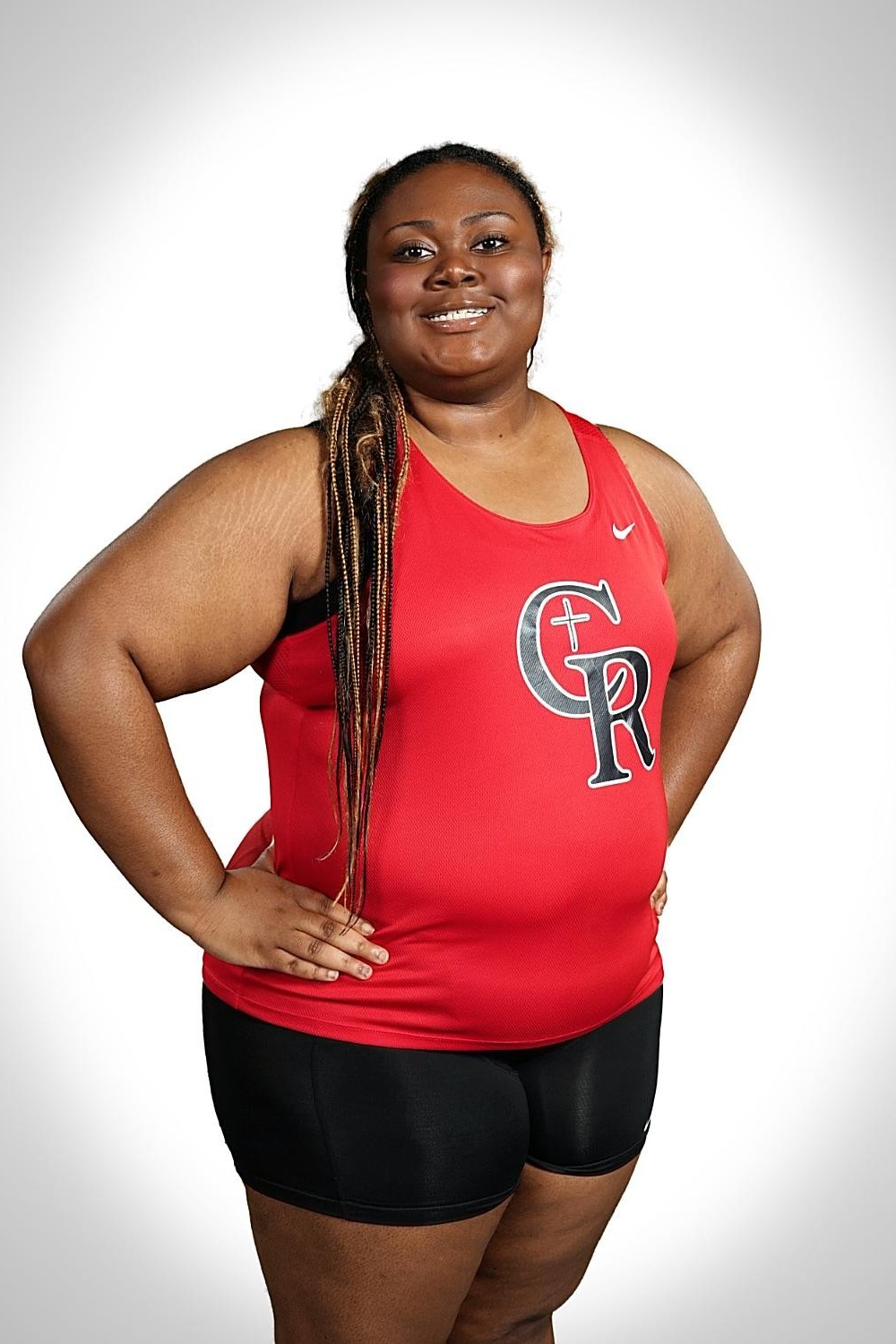 An adult woman with braids smiles and poses with her hands on her hips, wearing a red tank top with GR logo and black shorts.