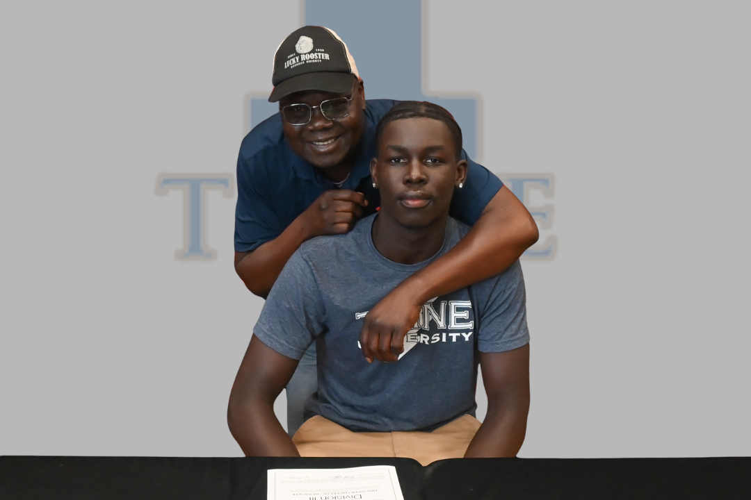 Two men sit behind a desk with the text "Signed Football" and "Orayo Urayo". One man in a cap stands behind the seated man.