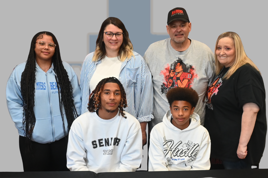 Group of people standing together behind a banner. One man and two boys wear white hoodies, one woman wears a blue hoodie, two women wear black shirts.