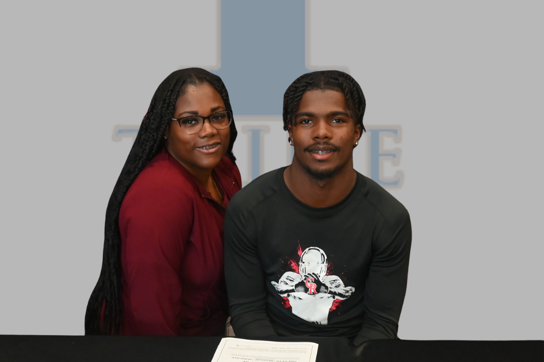 Two individuals sit behind a table, smiling. A man wears a black shirt with a graphic. A woman wears a red long-sleeved top and glasses.