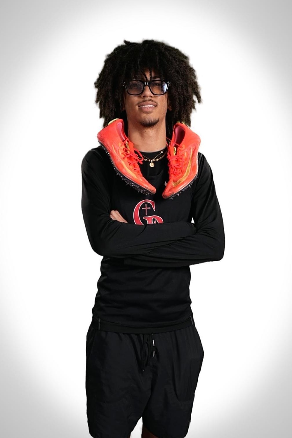 Person with curly hair and glasses poses in black outfit, folded arms, orange sneakers on shoulders.