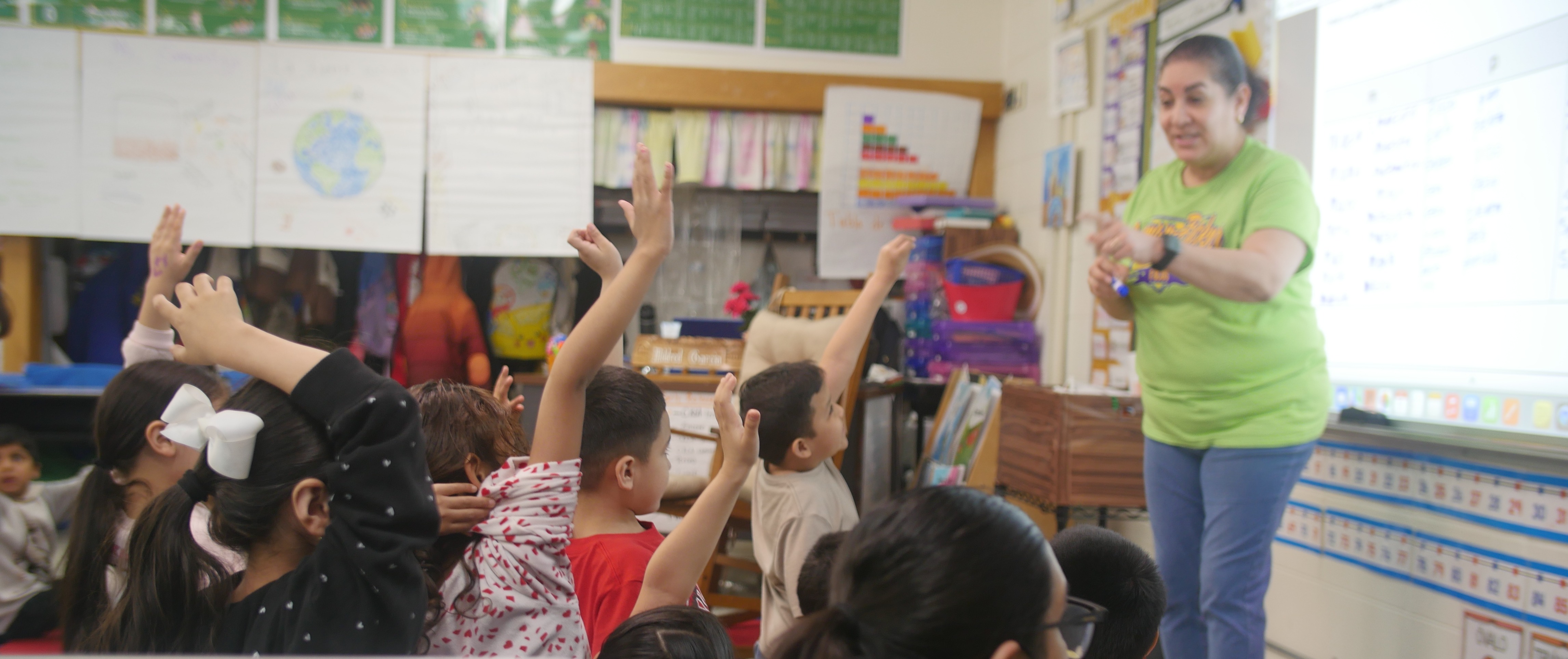 Students raise their hands to answer a question as a teacher presents a lesson on a projector screen