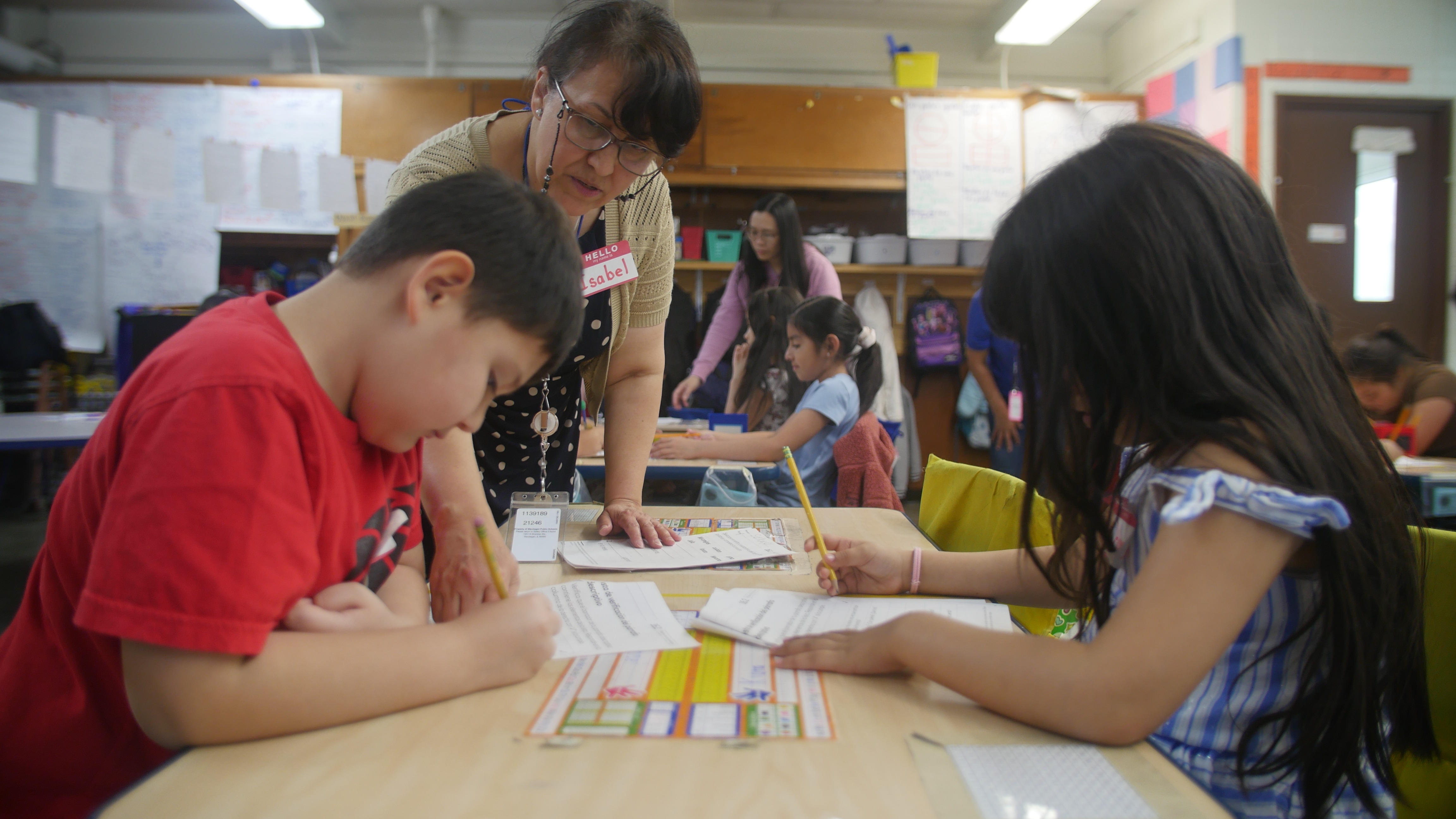 Students sit at grouped tables, holding pencils and focused on working through their classroom worksheets