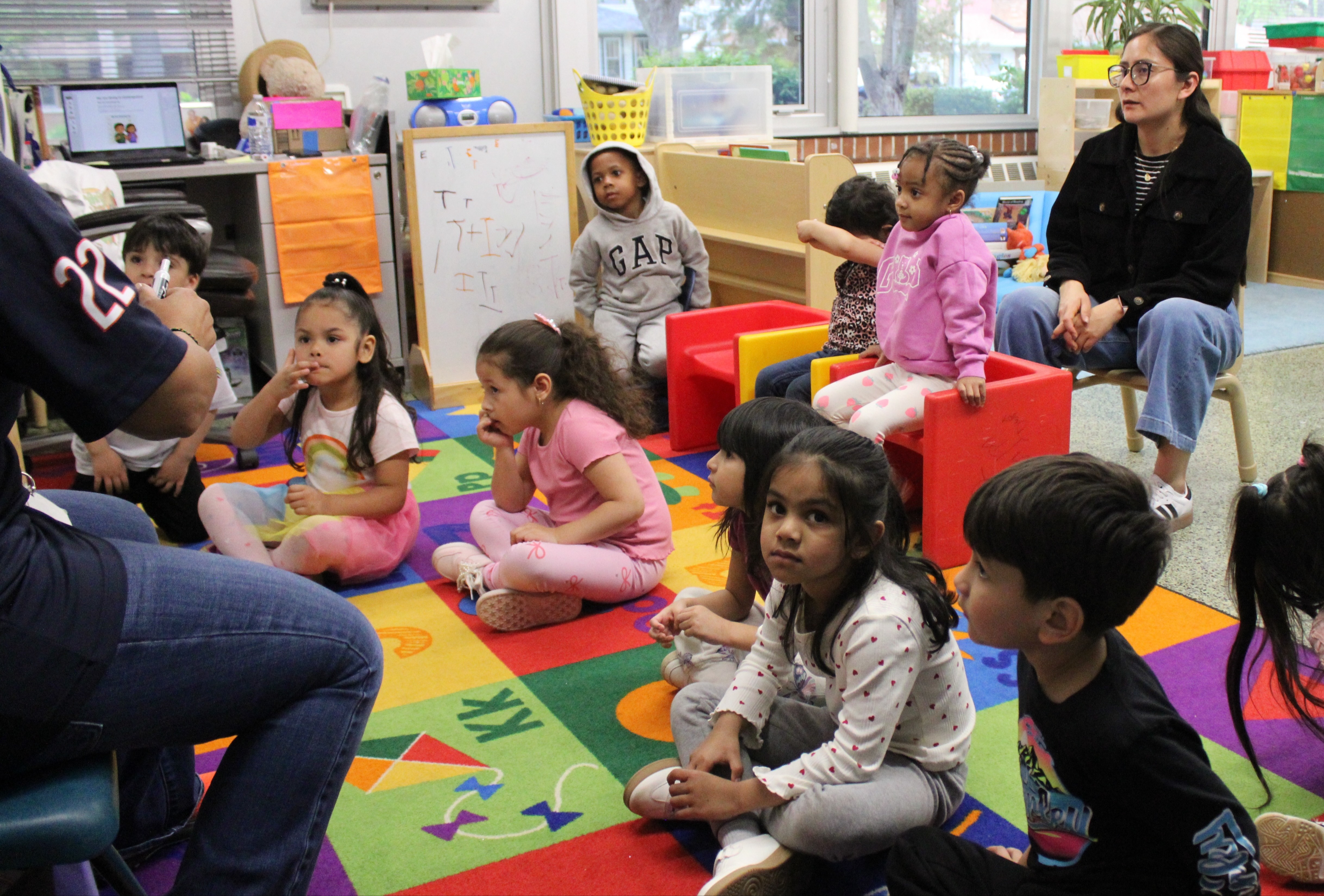 Students sit together in a group on the classroom floor, closely paying attention to their teacher.